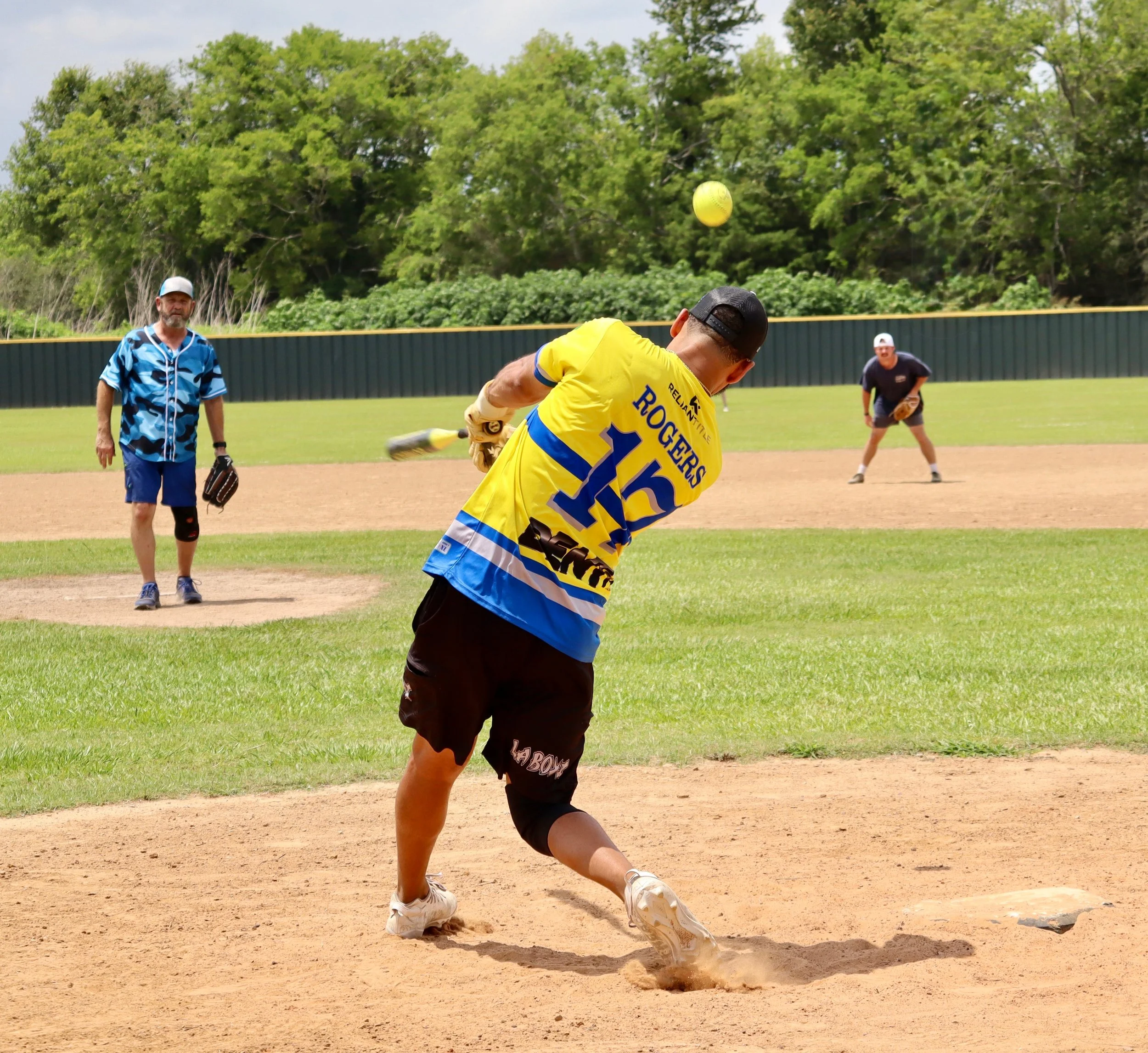 A man in a yellow and blue hockey jersey swings a baseball bat at a ball on a baseball field. Two other men are in the background, one standing near the pitcher's mound and the other ready in the outfield. The scene takes place outdoors on a sunny da
