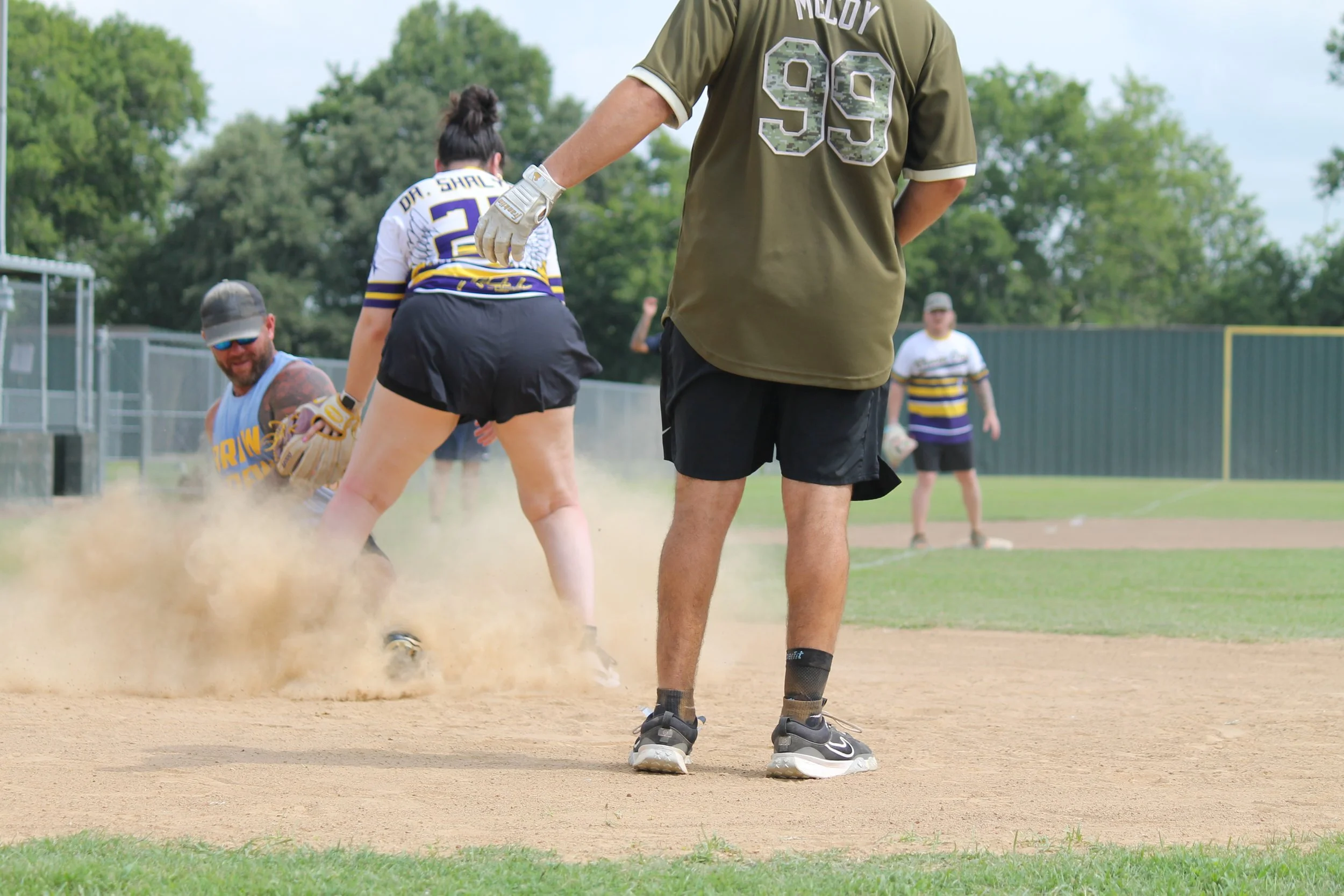 People playing softball on a field, with a woman sliding into a base as a man attempts to tag her, while a man in the background watches.