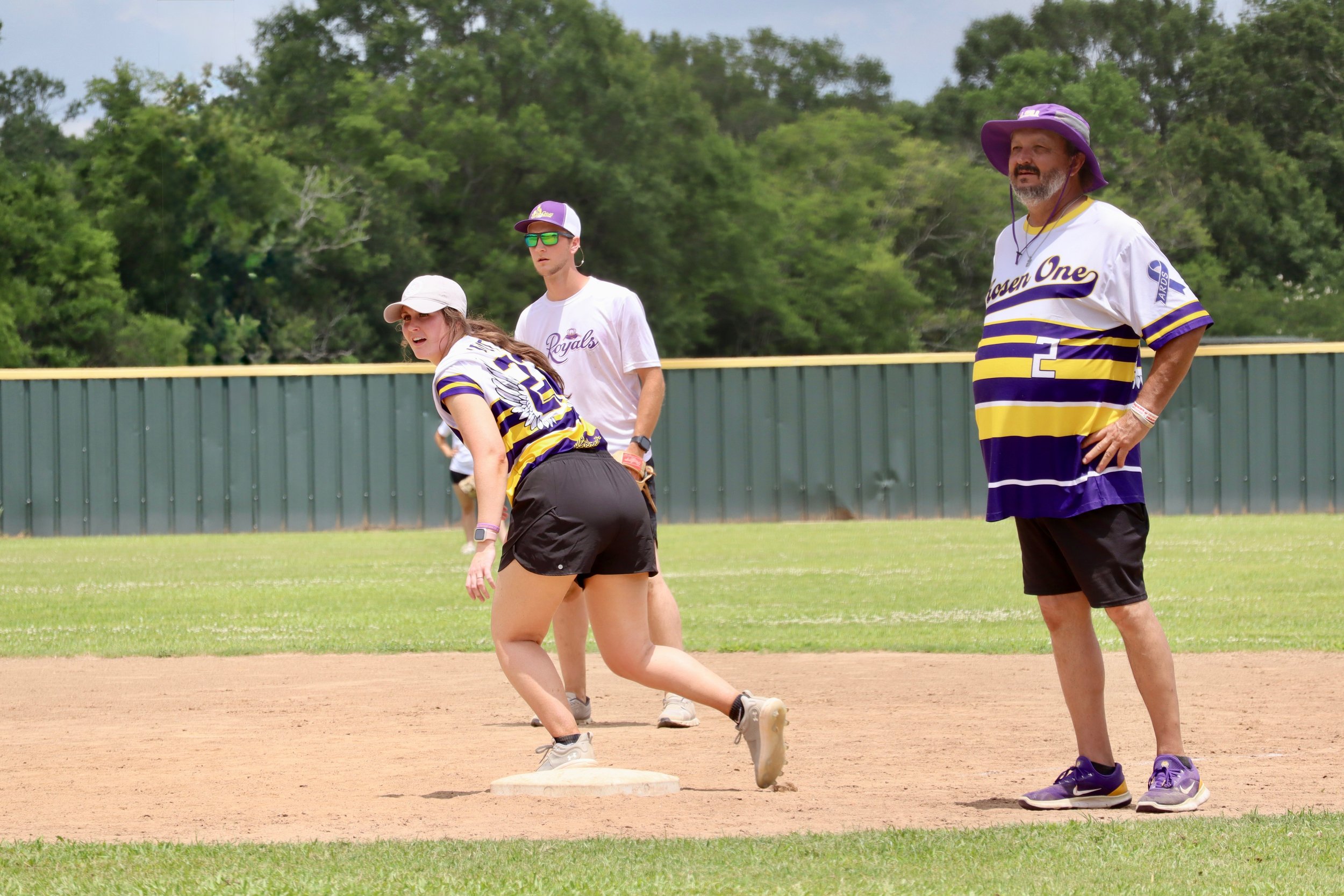 A group of people playing baseball on a field, with a girl preparing to run on a base, a man wearing a large purple hat and a Royals jersey standing on the side, and two young men in athletic gear in the background.