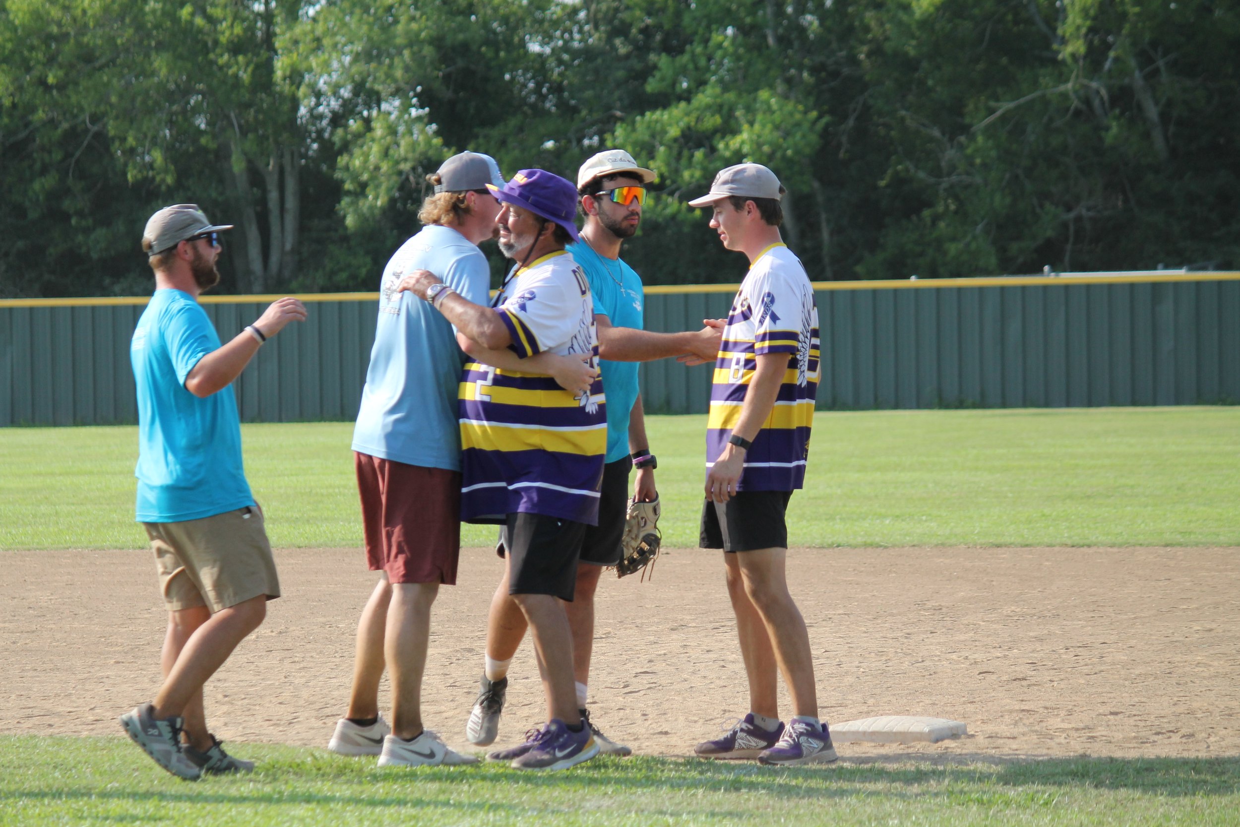 A group of five men on a baseball field, two embracing and shaking hands near a base, with lush green trees in the background.