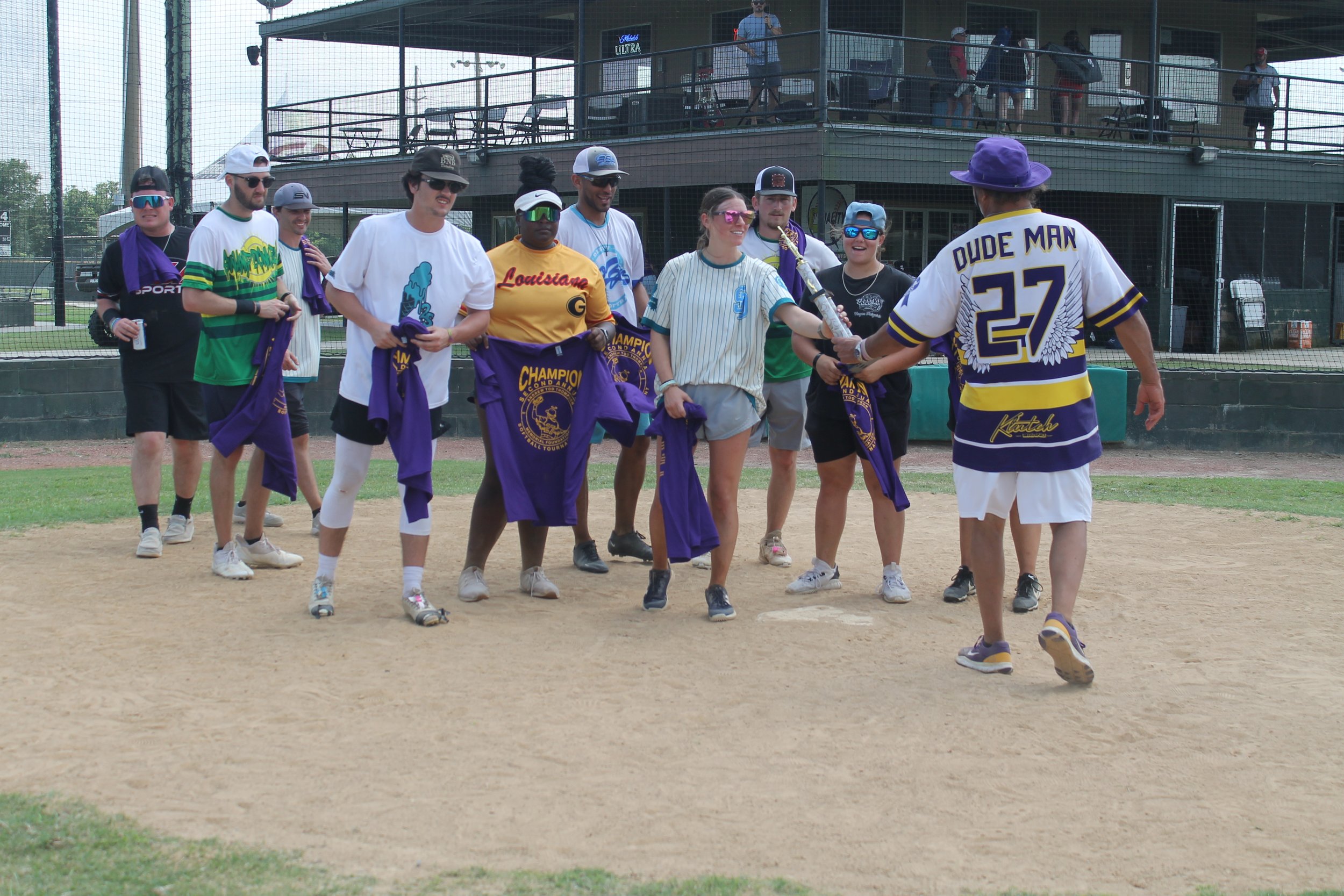 Group of people on a baseball field, some holding purple jackets, with a man in a sports jersey handing something to a young woman. Spectators are in the background on a second-story balcony.