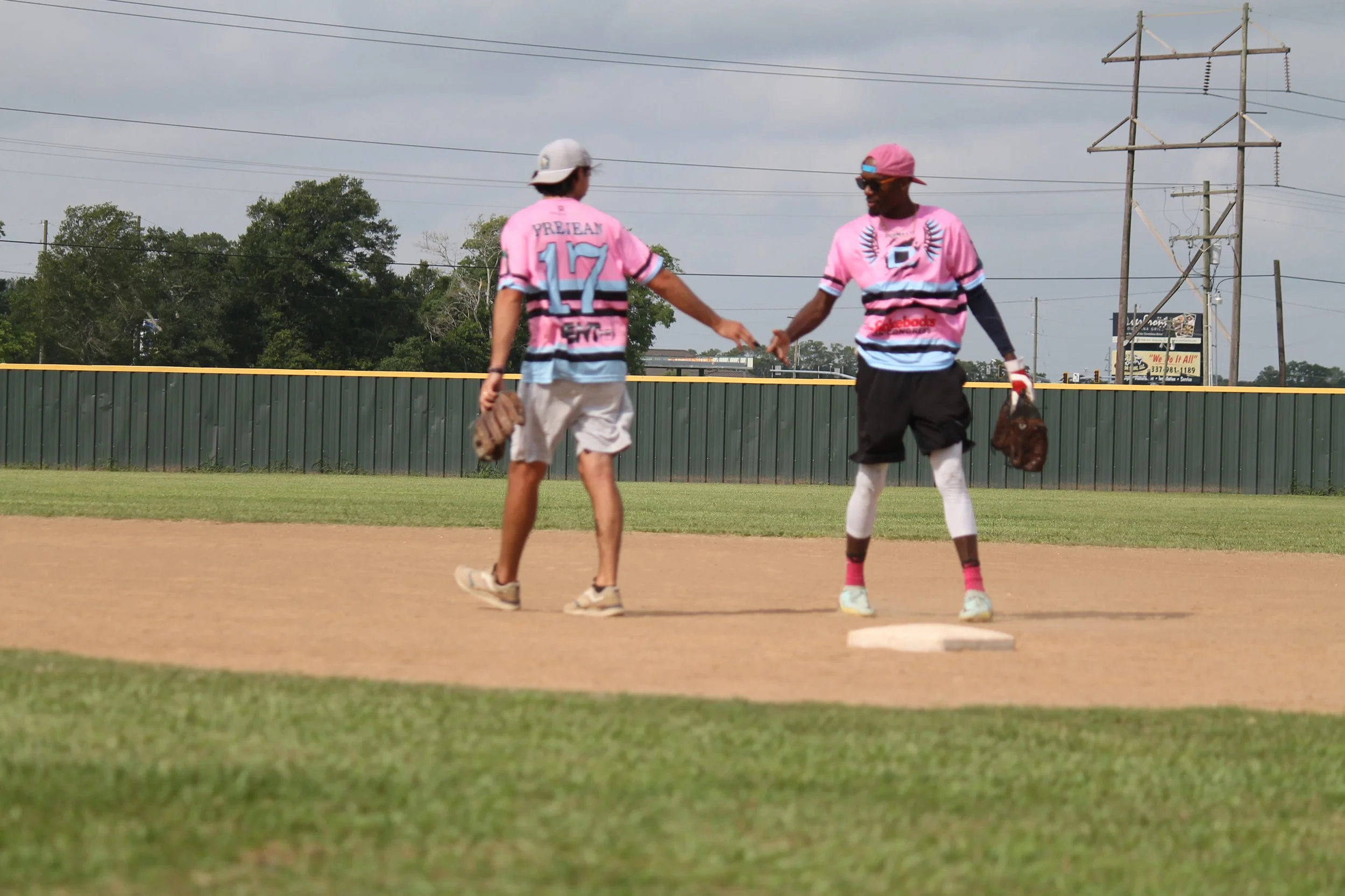 Two baseball players in pink uniforms shaking hands on the field, with a green fence and power lines in the background.