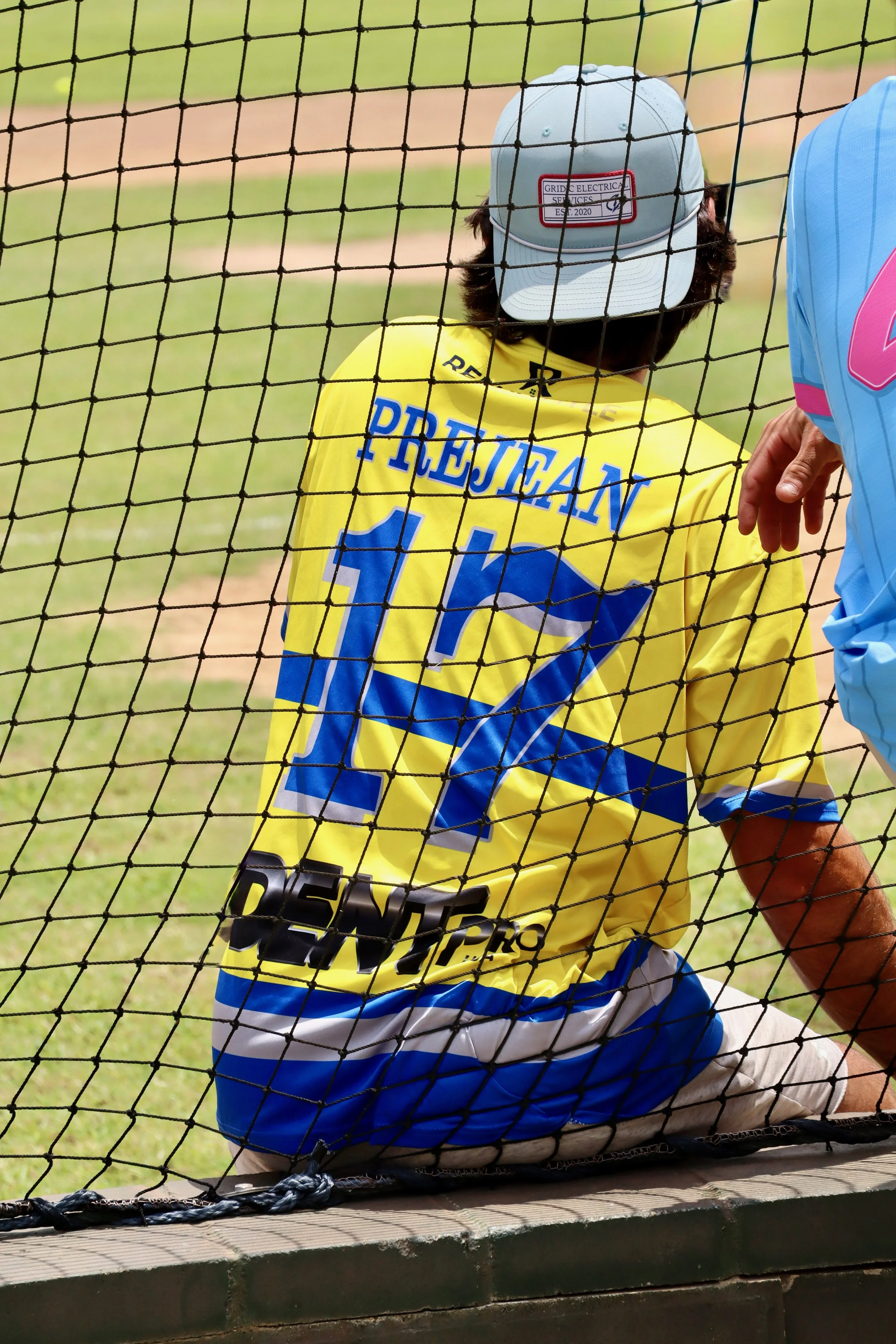 A person sitting on a bench at a sports field, wearing a yellow sports jersey with the name 'Prejean' and the number 17 on the back, and a gray cap. The picture is taken from behind a protective net.