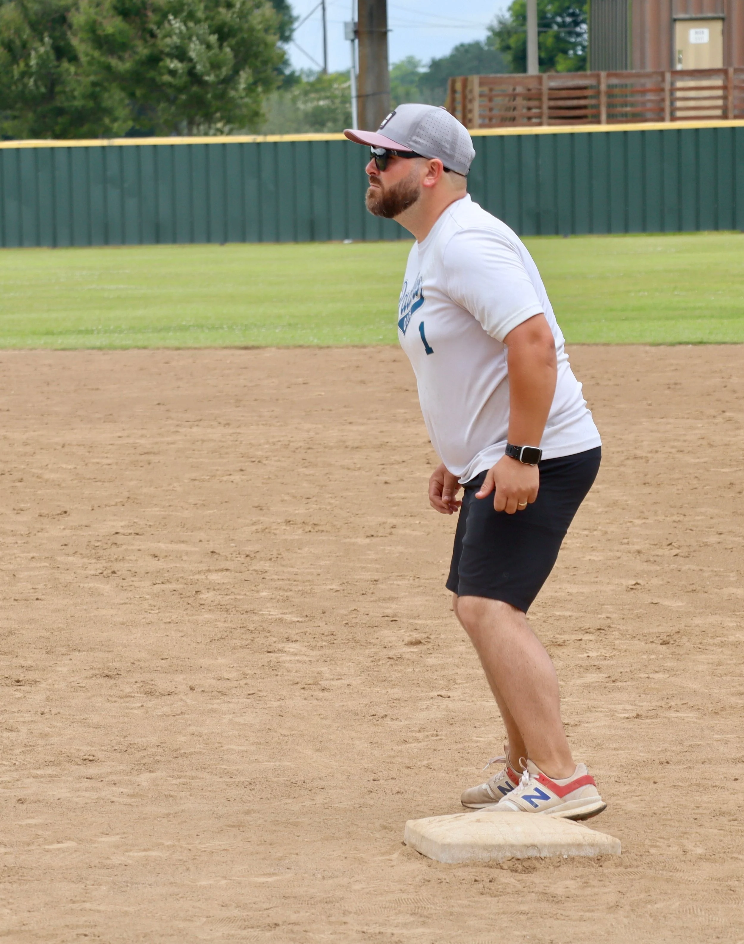 A man standing on a baseball field near a base, wearing sunglasses, a baseball cap, a smartwatch, a white t-shirt, black shorts, and sneakers, with a green fence and trees in the background.