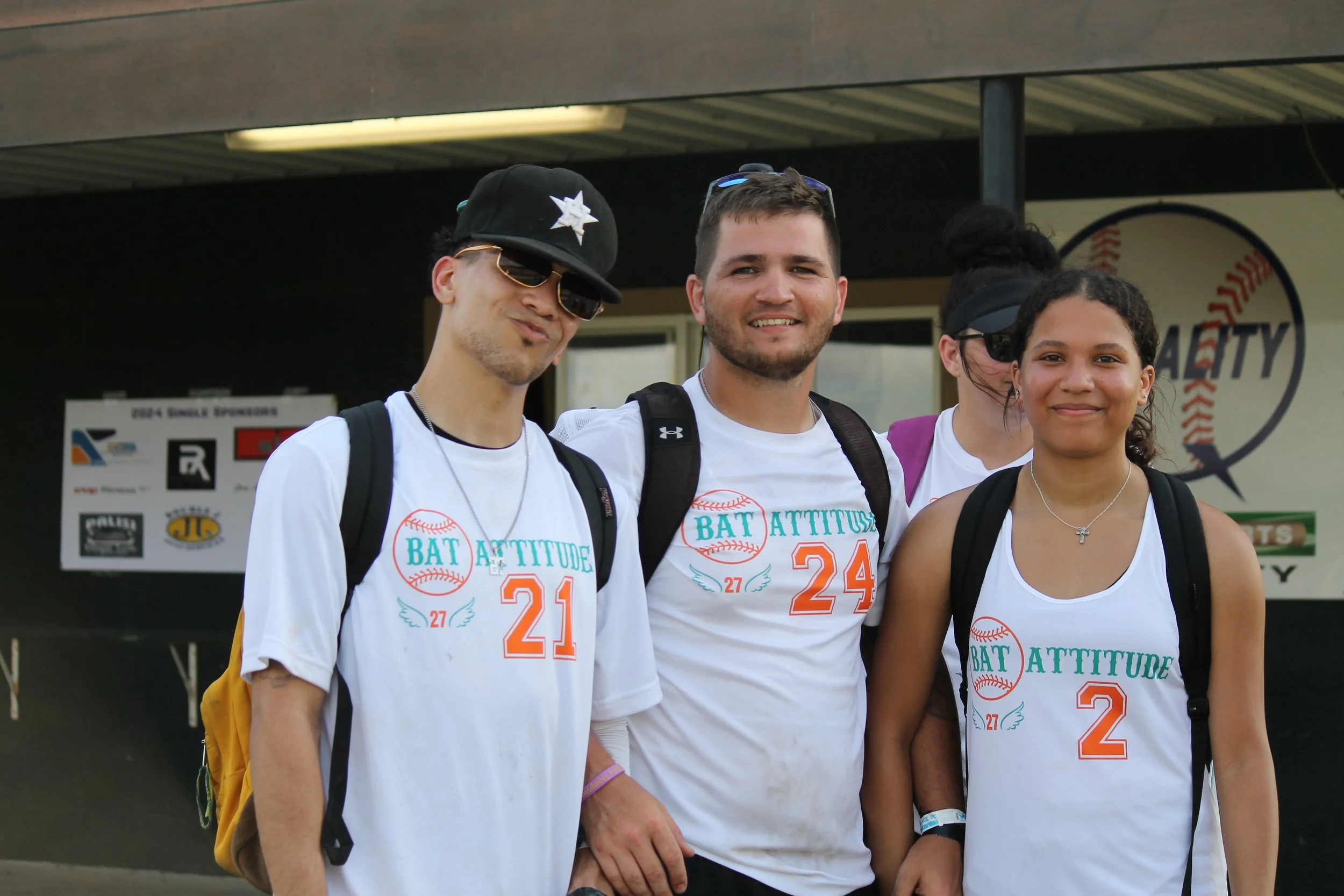 Group of four young people wearing white sports jerseys with 'Bat Attitude' written on them, smiling outdoors.