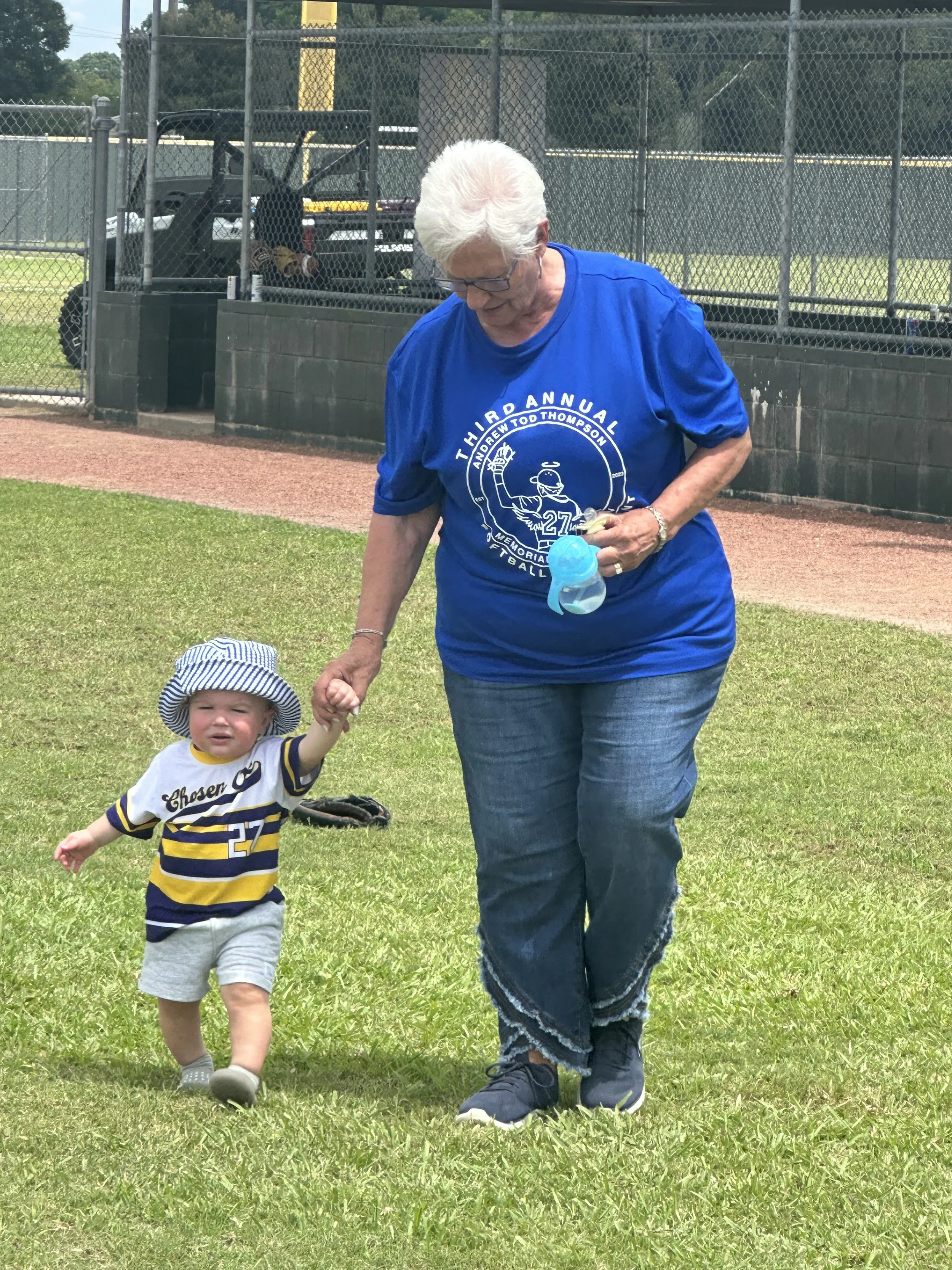 An older woman with white hair and glasses, wearing a blue t-shirt, holding hands with a small child wearing a striped shirt and hat, walking on grass near a baseball field.