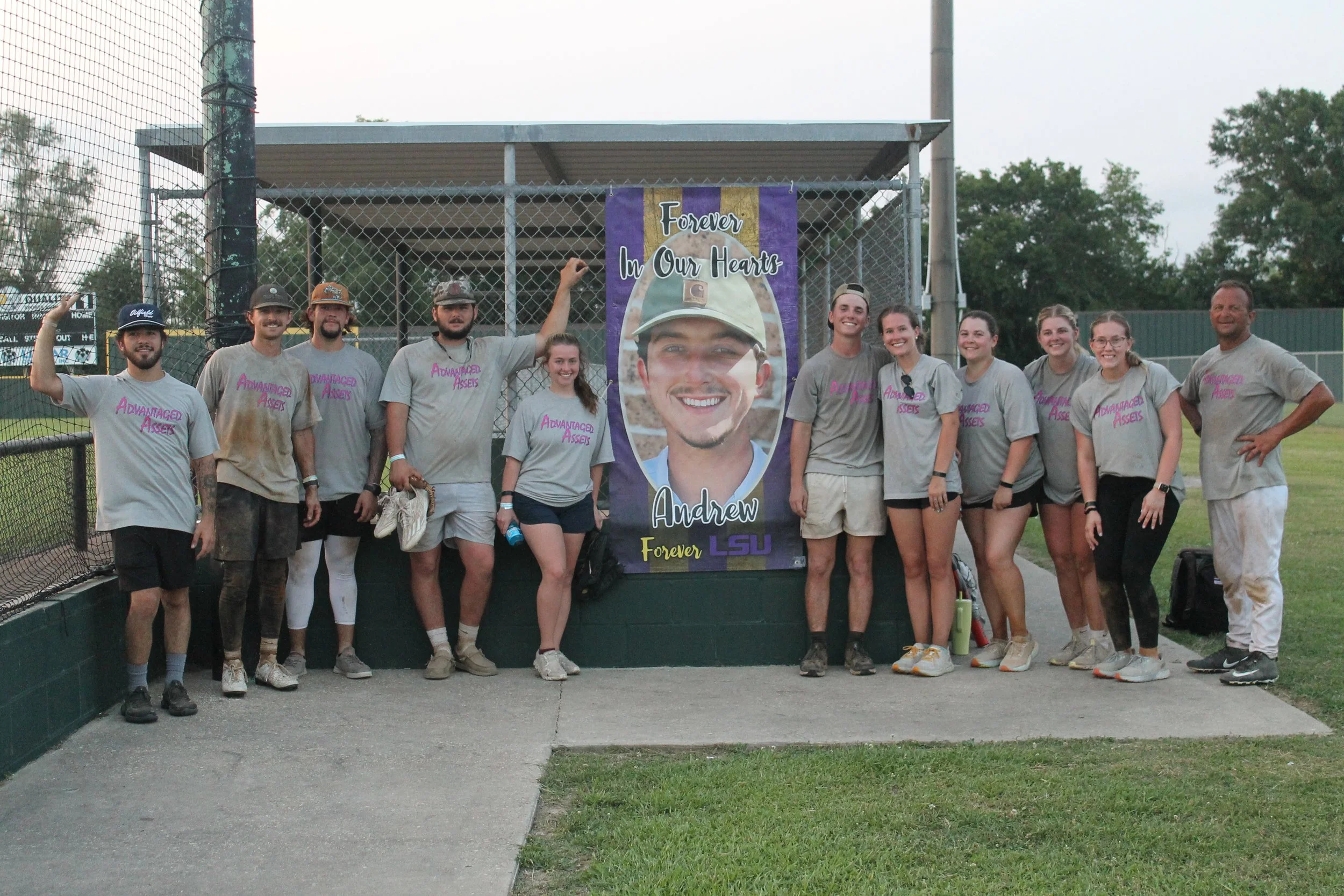 Group of people at a baseball field standing in front of a large photo of a smiling young man and a banner that reads 'Forever In Our Hearts, Andrew, Forever, LSU.' The group is wearing matching gray shirts with pink text 'Advantaged Assets' and is s