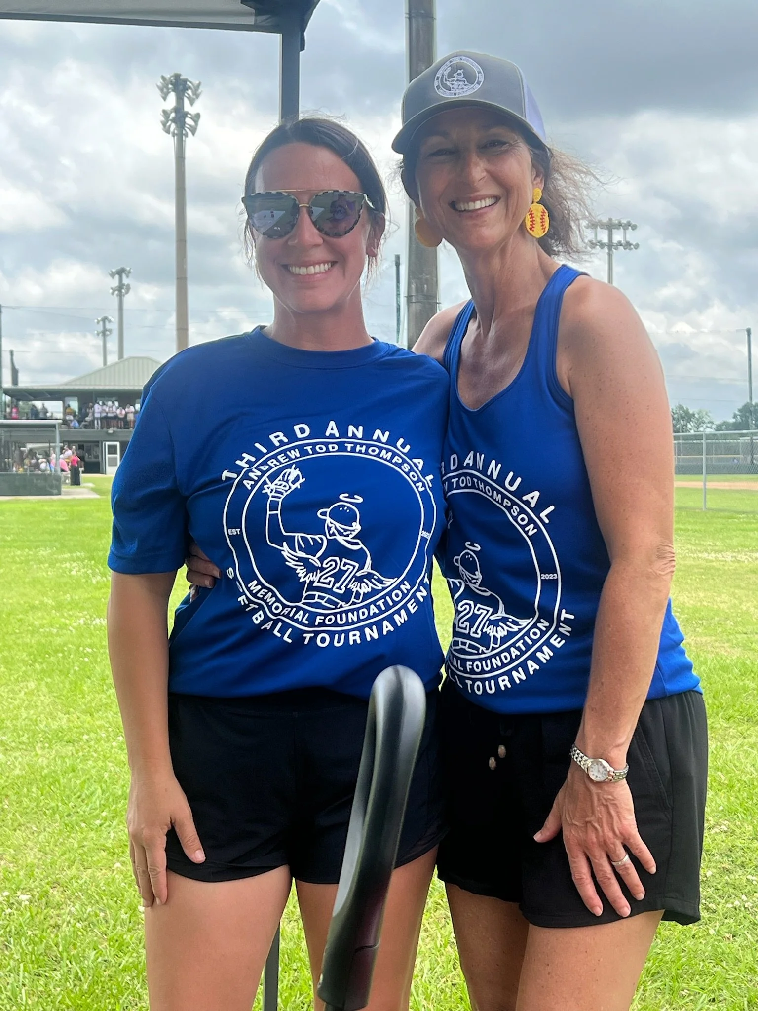 Two women wearing blue T-shirts with 'Andrew Todd Thompson Memorial Foundation' and a bird logo, standing on a baseball field. They are smiling, with one wearing sunglasses and the other wearing a cap and earrings, and the background shows a baseball