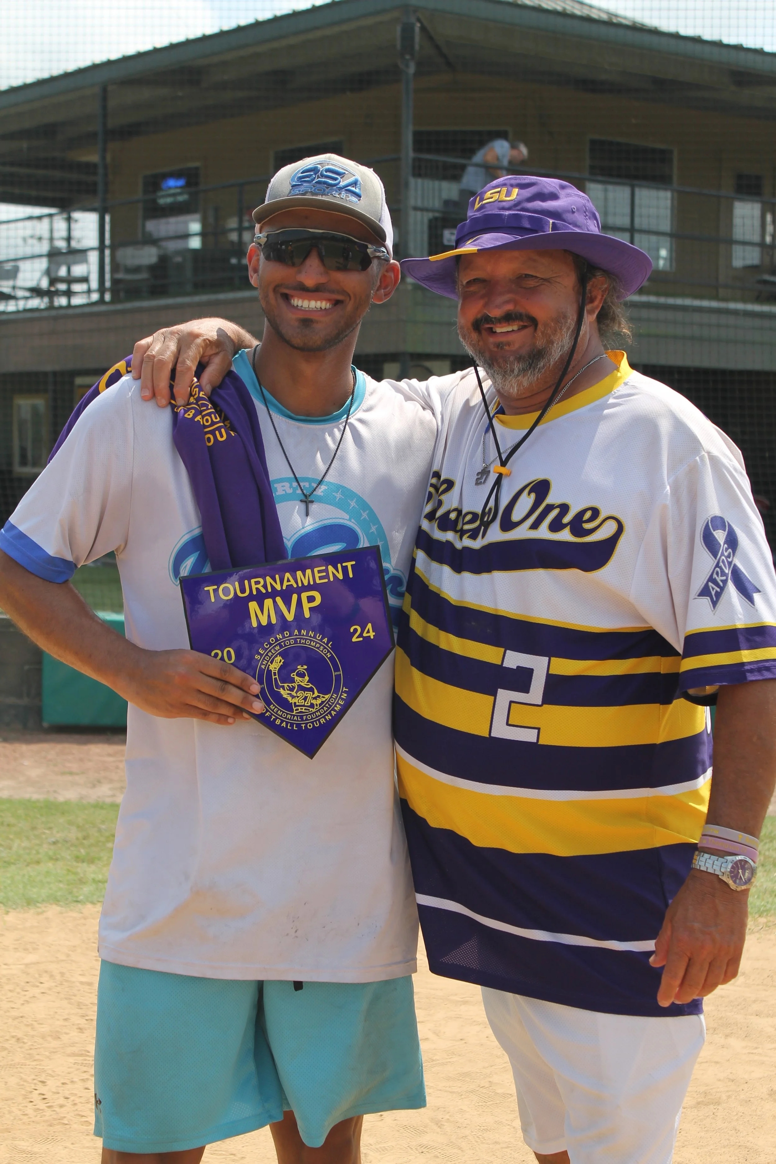 Two men smiling and standing close together on a baseball field. The man on the left is holding an MVP tournament plaque. The man on the right is wearing a LSU hat and jersey with the number 2. Both are dressed casually, and in the background, there 