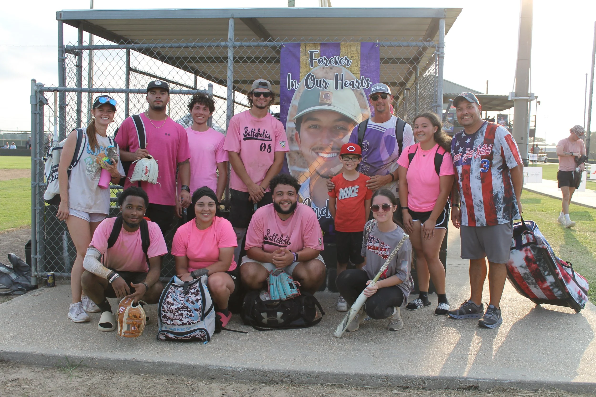 A group of people, including children and adults, gathered on a baseball field in front of a chain-link fence with a large poster of a smiling young man in military uniform. Some are wearing pink shirts supporting awareness, and others have backpacks