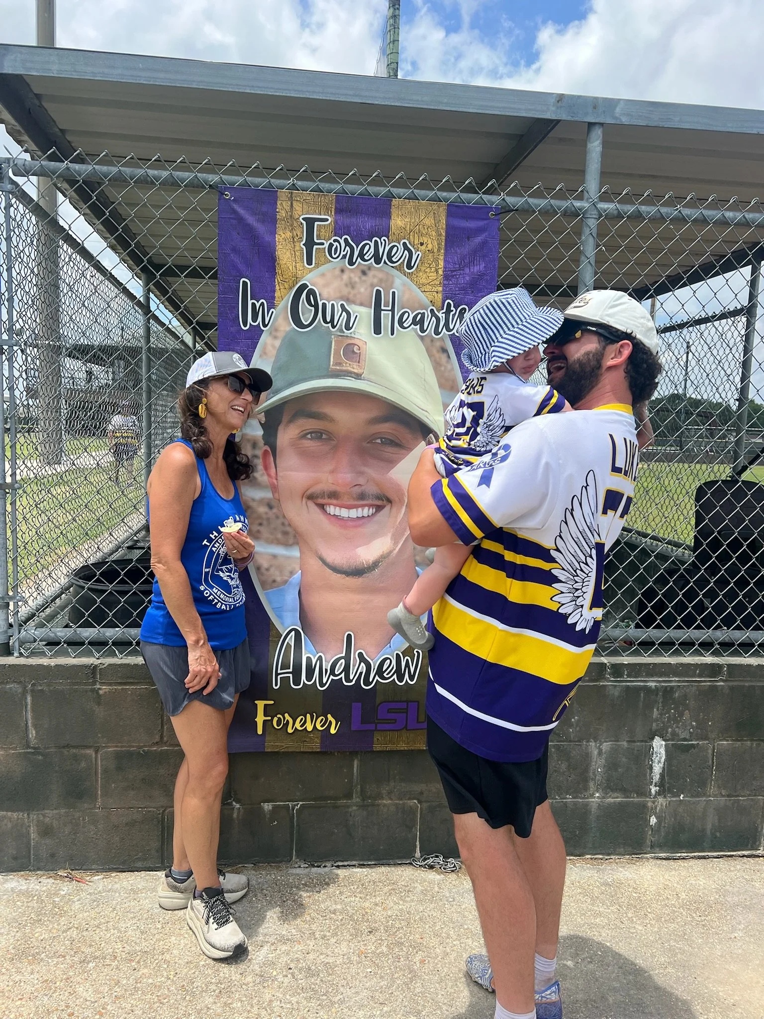 A woman and a man holding a young child near a large memorial banner with a smiling young man in a military helmet. The banner has the words "Forever In Our Heart" and "Andrew." The woman is wearing a blue shirt, gray shorts, a white cap, and sunglas