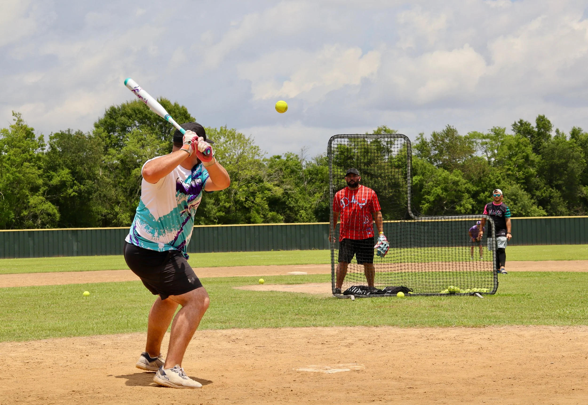 A person at bat practicing softball in the batting cage, with a pitcher and another person observing on the field.