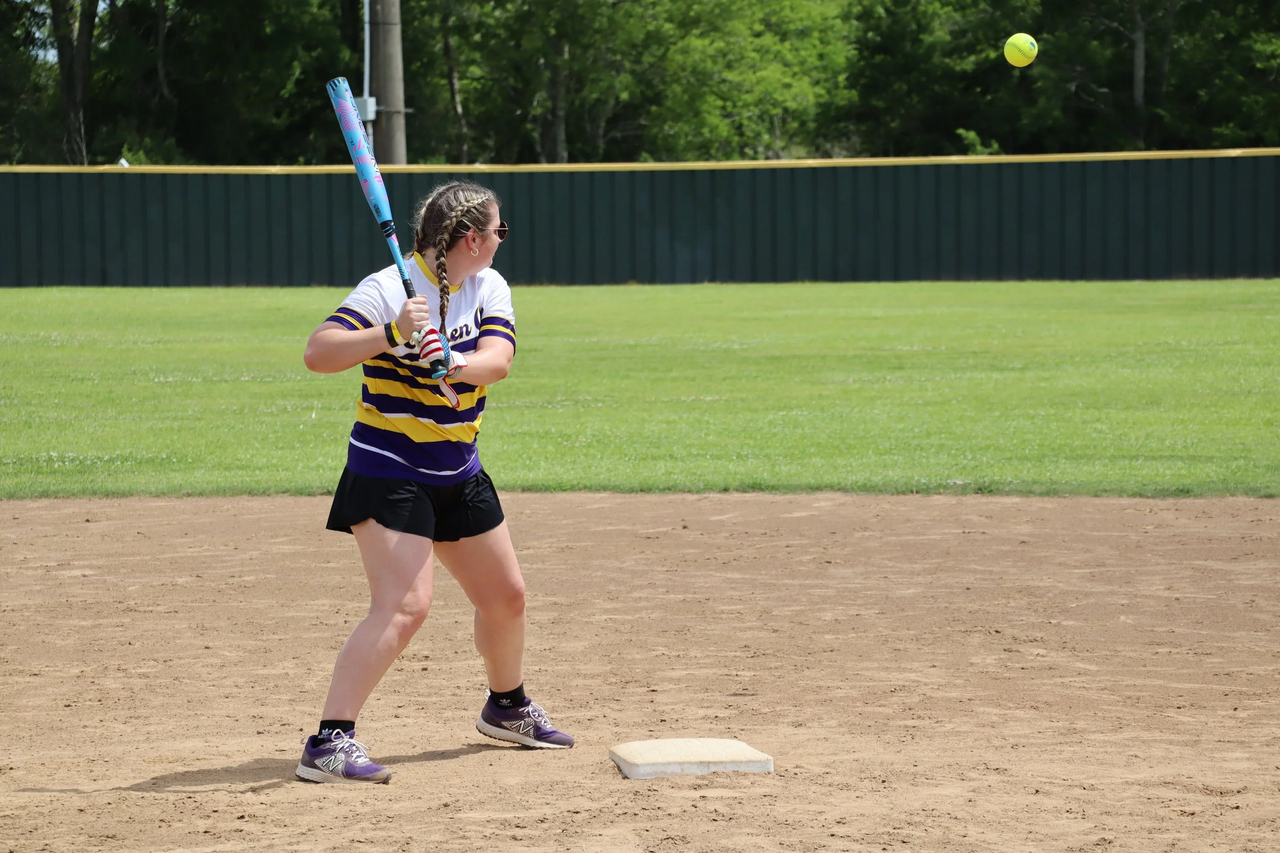 A woman playing softball, preparing to hit the ball at a base on the dirt field, with green grass and trees in the background.
