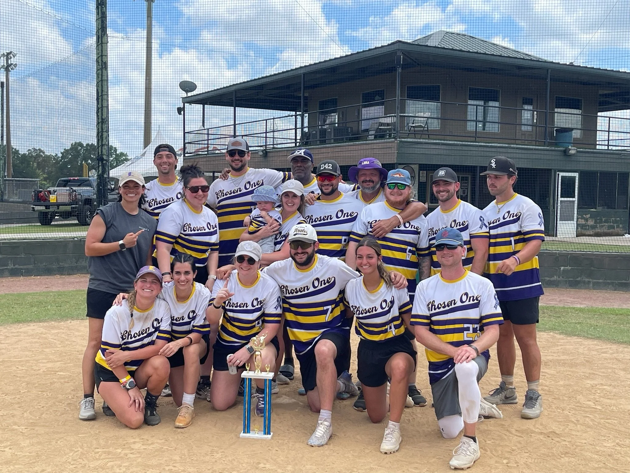 Group of people in baseball uniforms on a field, holding a trophy, posing for a photo, with a building and cloudy sky in the background.