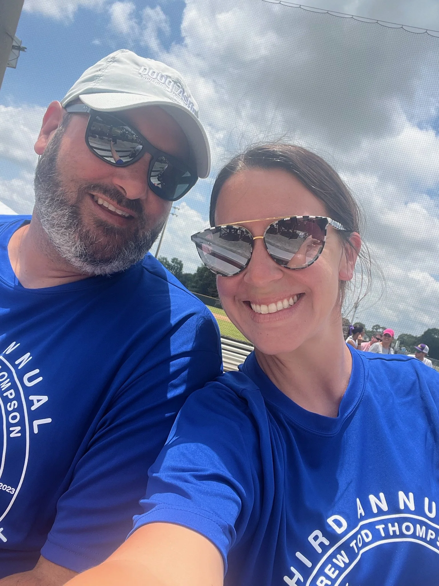 Two smiling people taking a selfie outdoors, both wearing blue shirts and sunglasses, with a cloudy sky and a baseball field in the background.