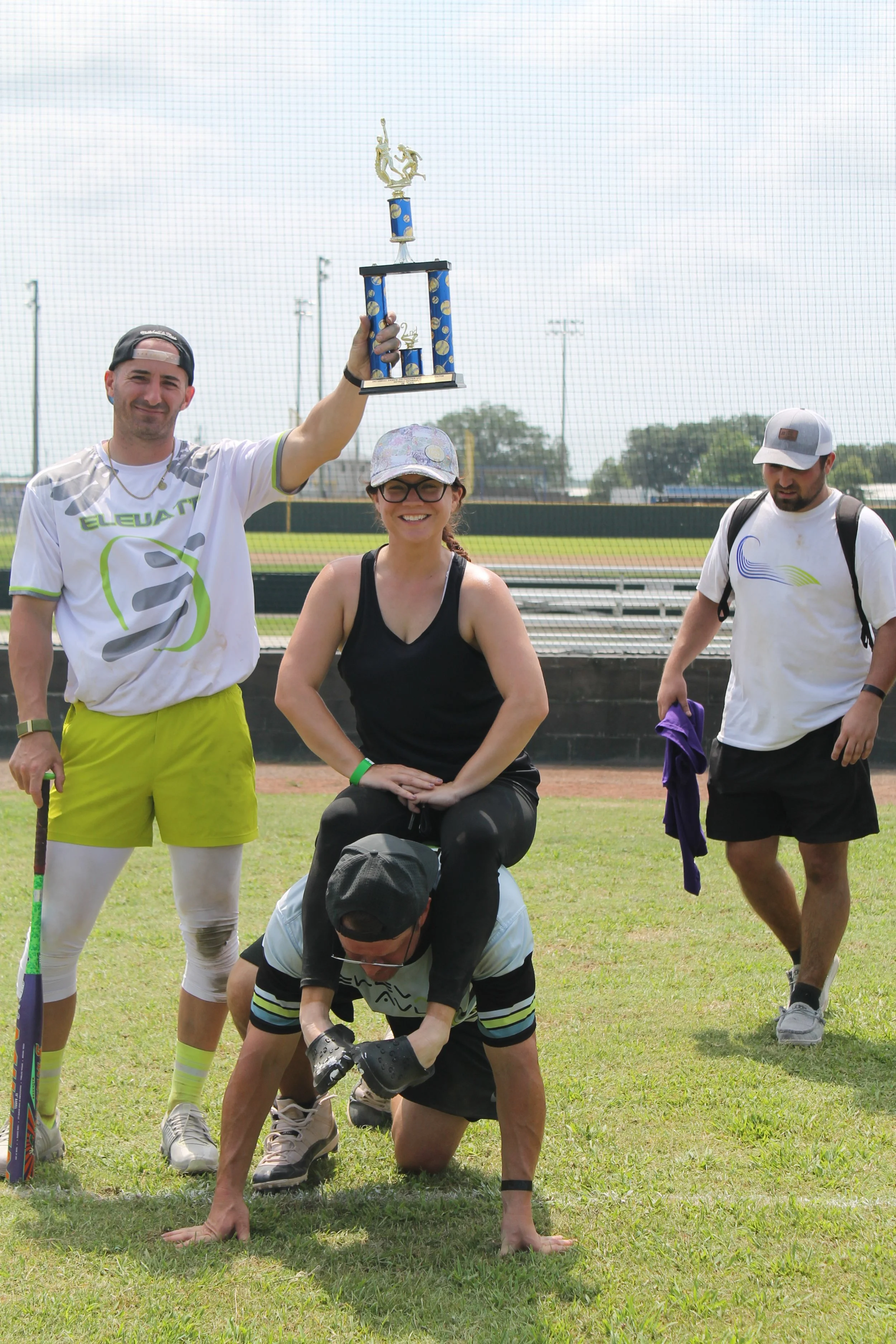 Group of four people celebrating a sports victory on a field; one man holds a trophy overhead, woman sits on a man's shoulders, and two other men stand nearby, one holding a rolled-up towel.