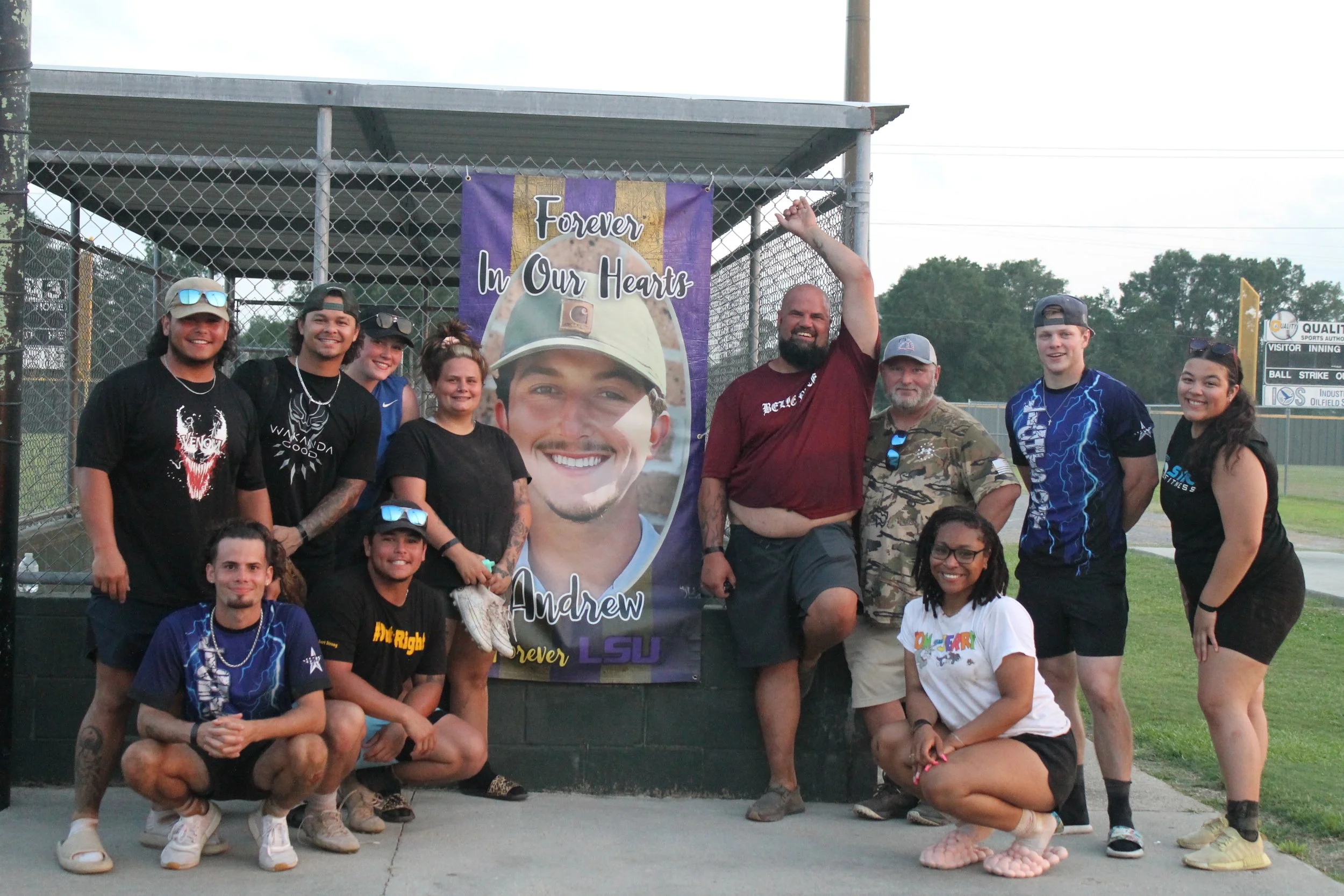 Group of people at a baseball field standing in front of a large banner that reads 'Forever in Our Hearts Andrew'. People are smiling, posing, and some have tattoos or sunglasses. The group includes both men and women of various ages.