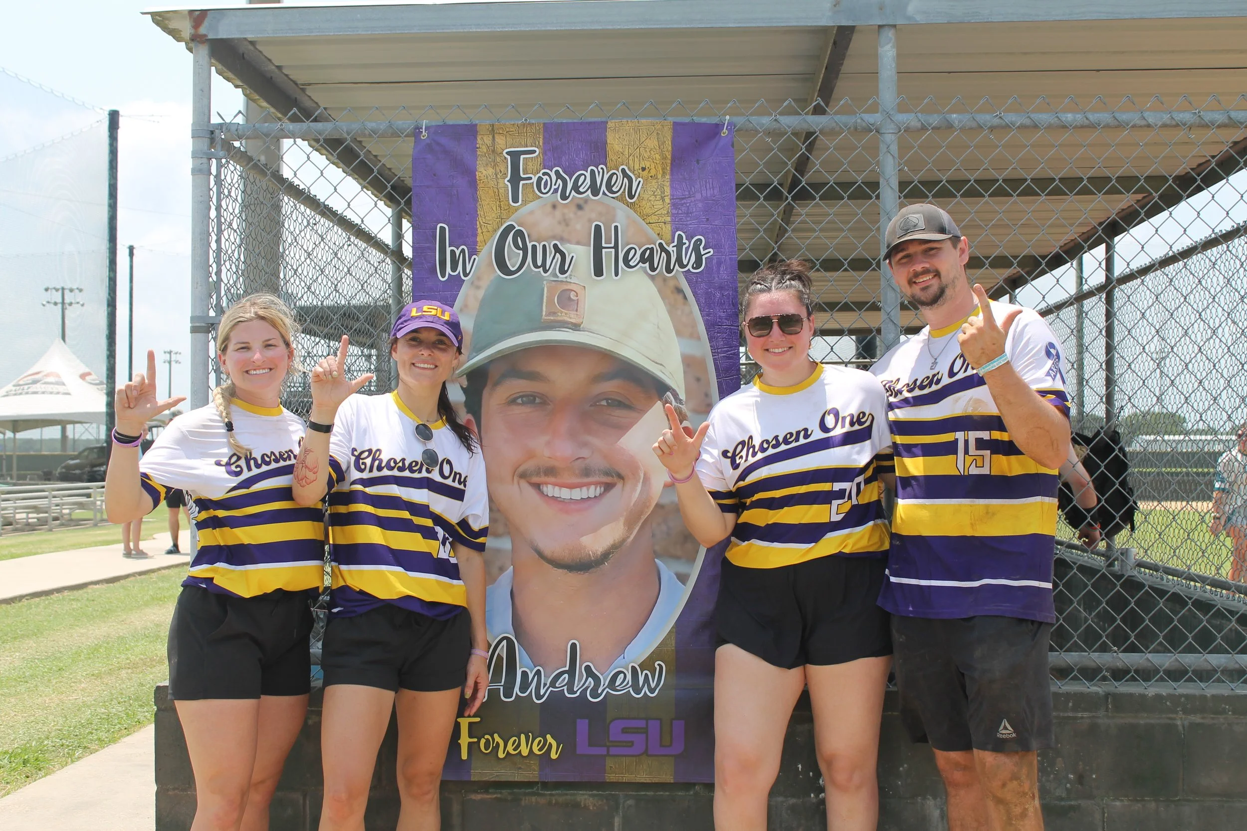 Four people in LSU sports jerseys standing outdoors in front of a large memorial banner with a photo of a smiling young man wearing a helmet, with the text ‘Forever In Our Hearts’ and ‘Andrew Forever LSU’ on it. They are making hand gestures and smil