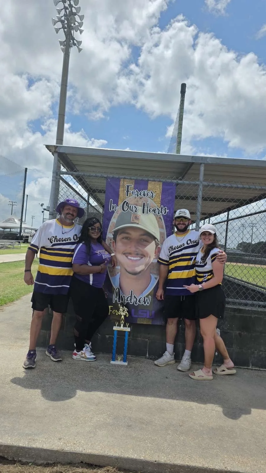 A group of five people posing in front of a large banner with a young man's portrait and the words "Forever In Our Hearts" and "Andrew." They are at a baseball field, some wearing purple and gold apparel with the LSU logo, and there is a trophy on th