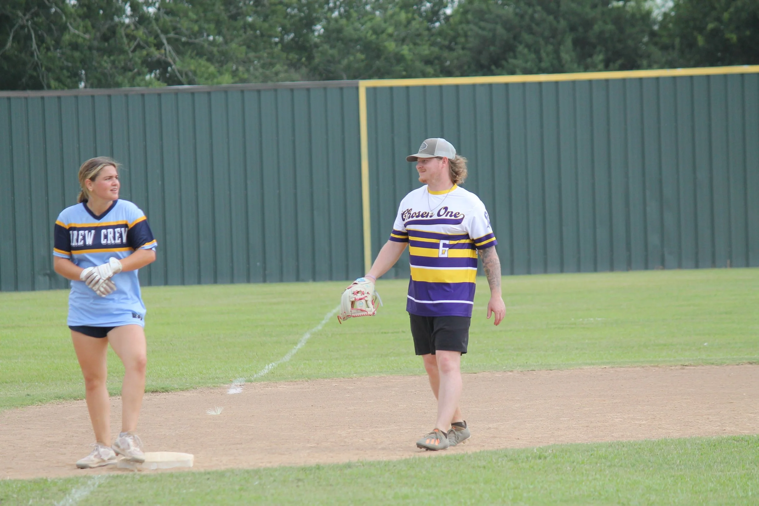 A woman wearing a blue softball uniform and gloves, standing on a base, talks to a man wearing a baseball jersey with purple and yellow stripes, black shorts, and a cap, watering the base on a grassy field with a green fence in the background.