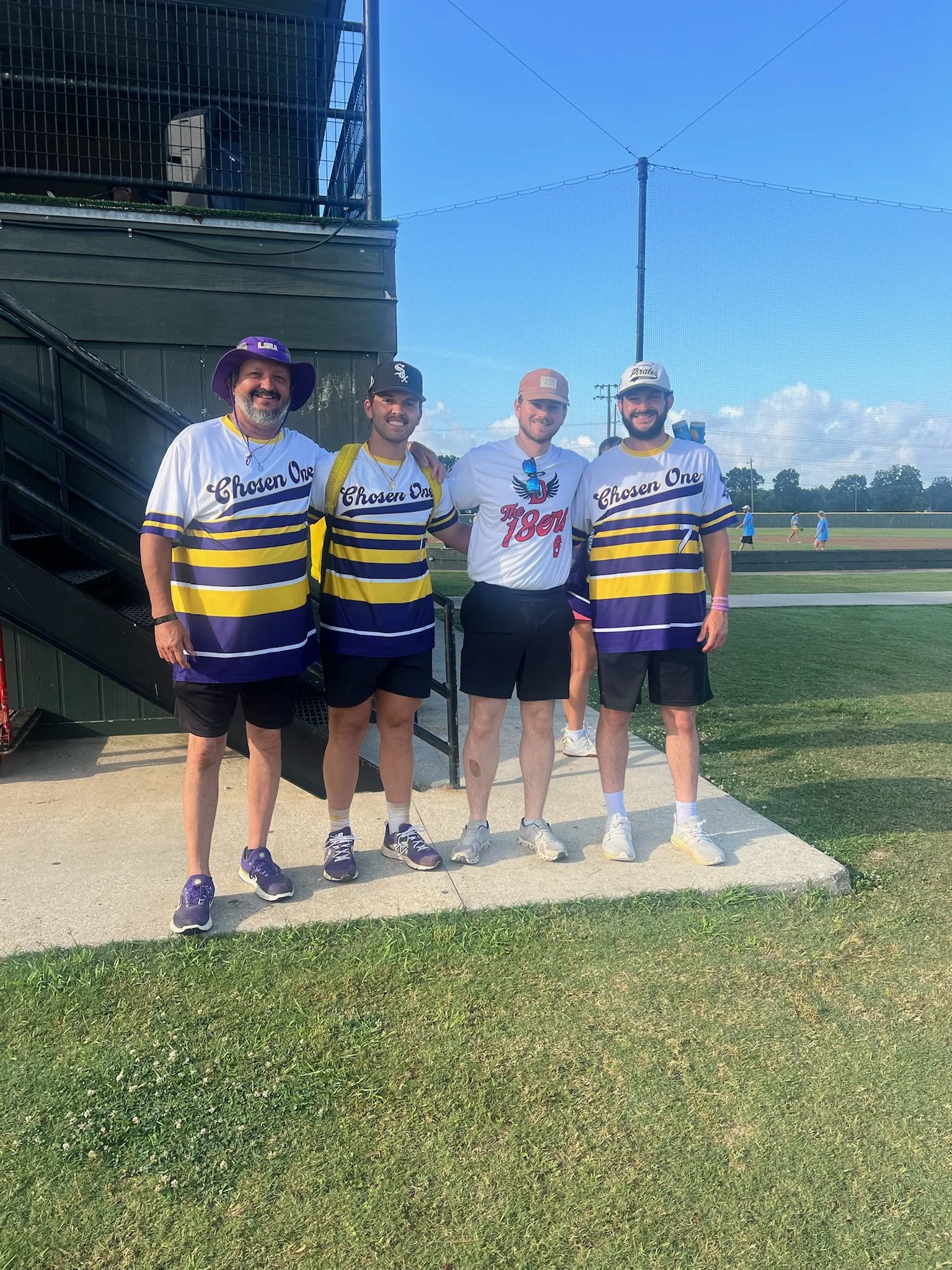 Four men standing together outdoors on a sunny day, wearing sports jerseys and baseball caps, with a baseball field in the background.