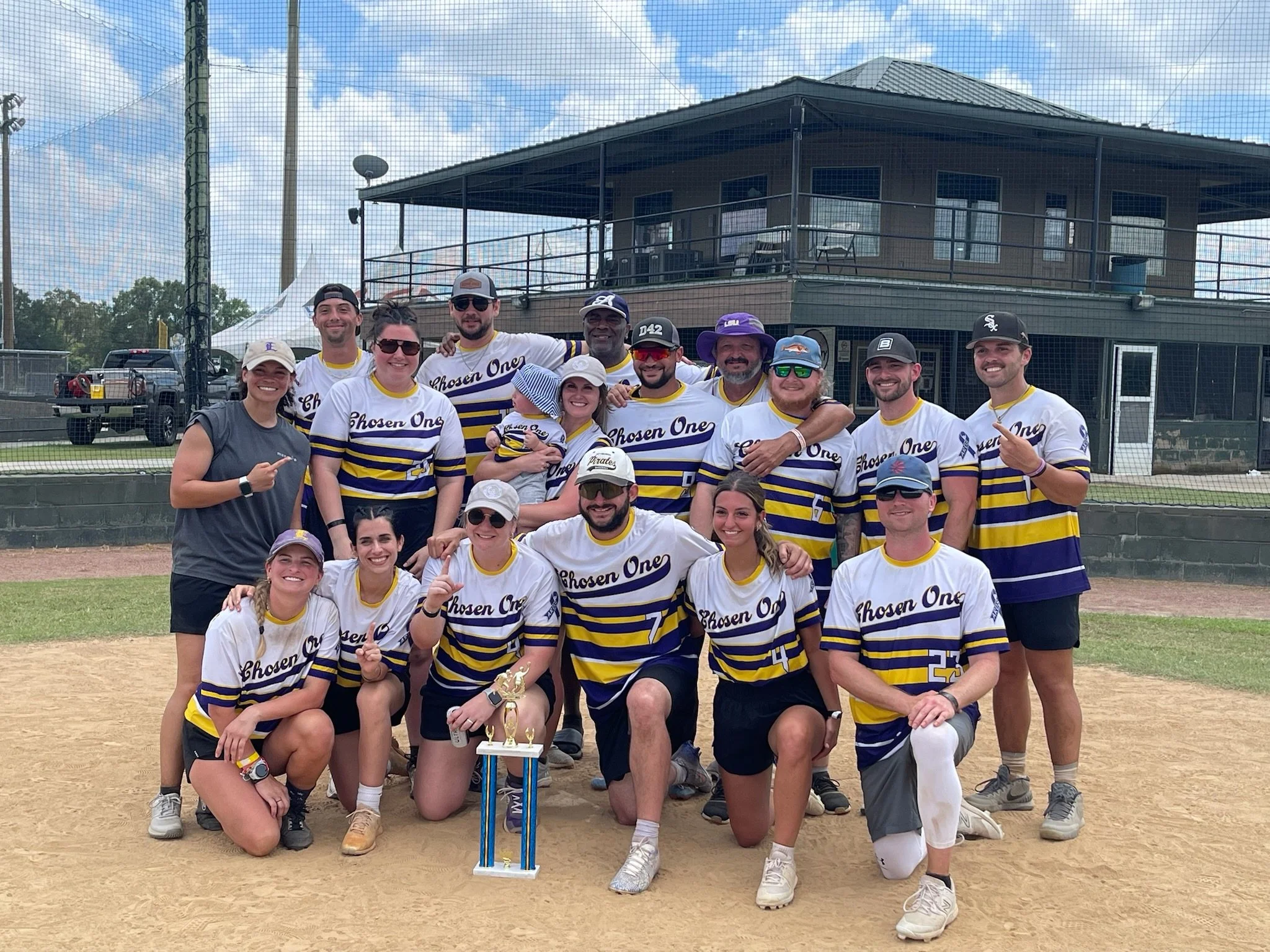 A group of people posing on a baseball field with a trophy, wearing matching jerseys that say 'Chosen One.' They are smiling and celebrating.