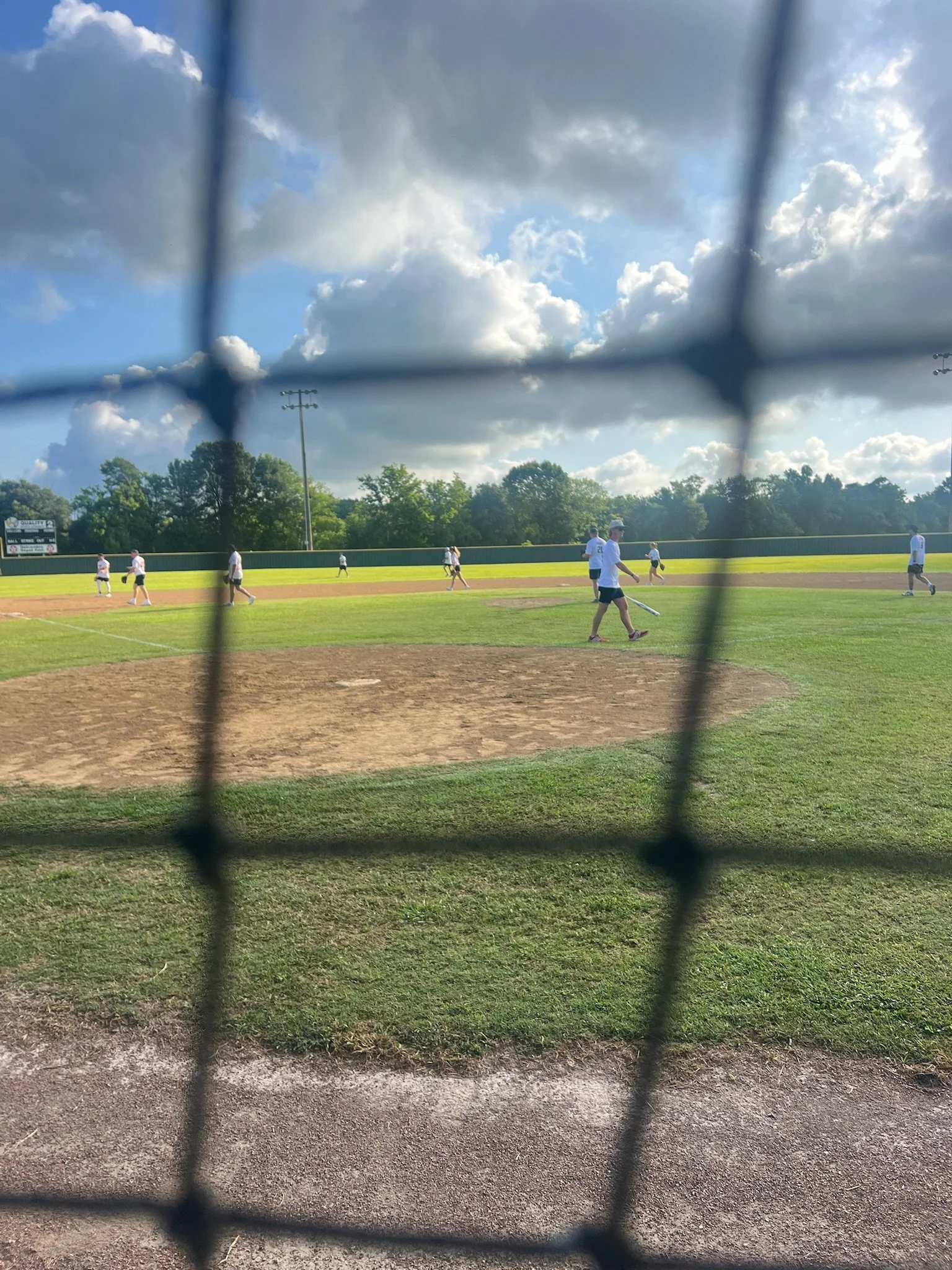 View of a baseball field seen through a chain-link fence with players practicing on the field on a partly cloudy day.