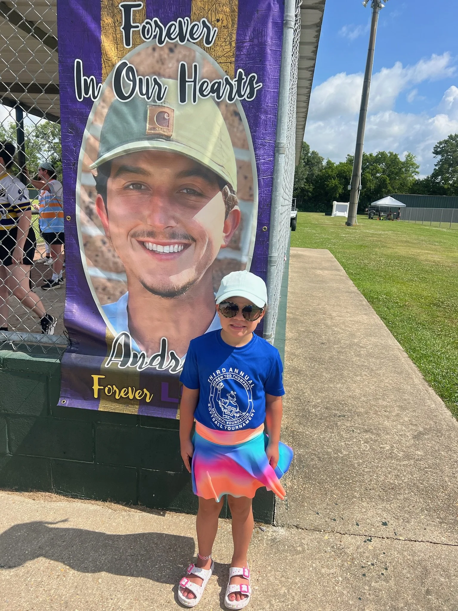 A young girl wearing sunglasses, a white cap, a blue T-shirt, and a colorful skirt standing outside next to a large banner that reads 'Forever in Our Hearts' with a smiling man's face on it.