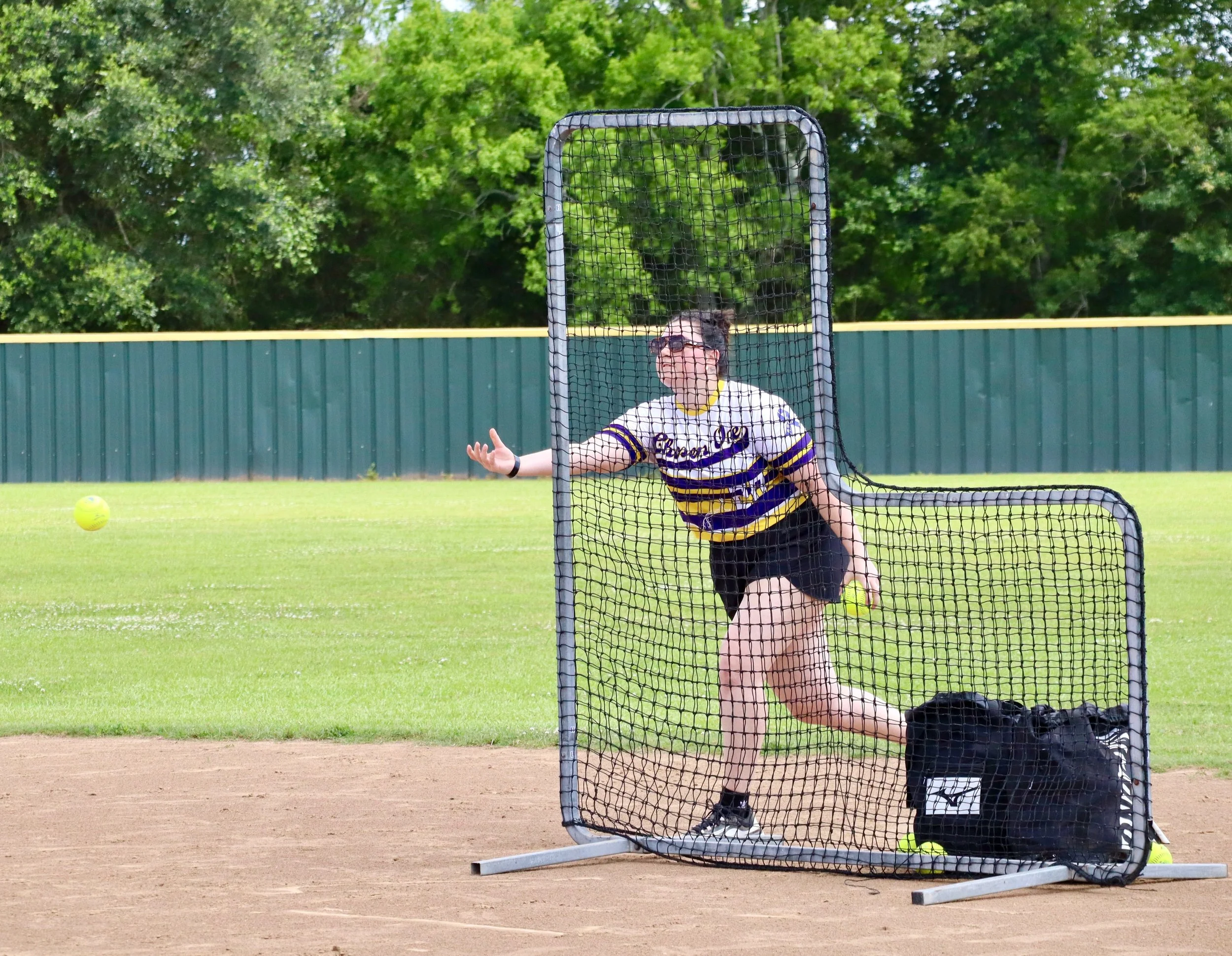 Person practicing softball batting in a batting cage on a field with green grass and trees in the background.