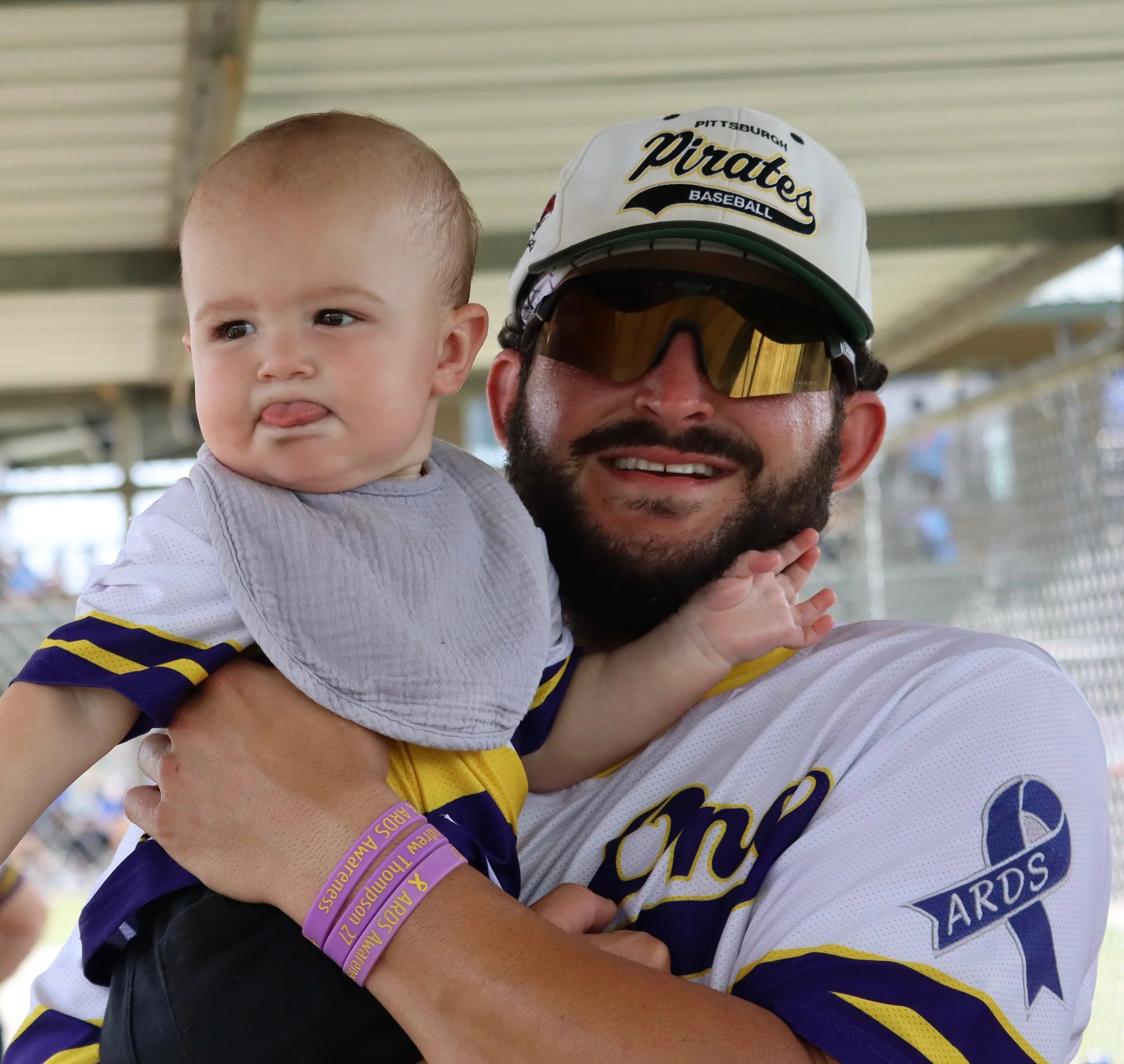A man holding a young child, both wearing sports jerseys. The man wears sunglasses and a baseball cap. They are at a sporting event, with a stadium and other spectators in the background.