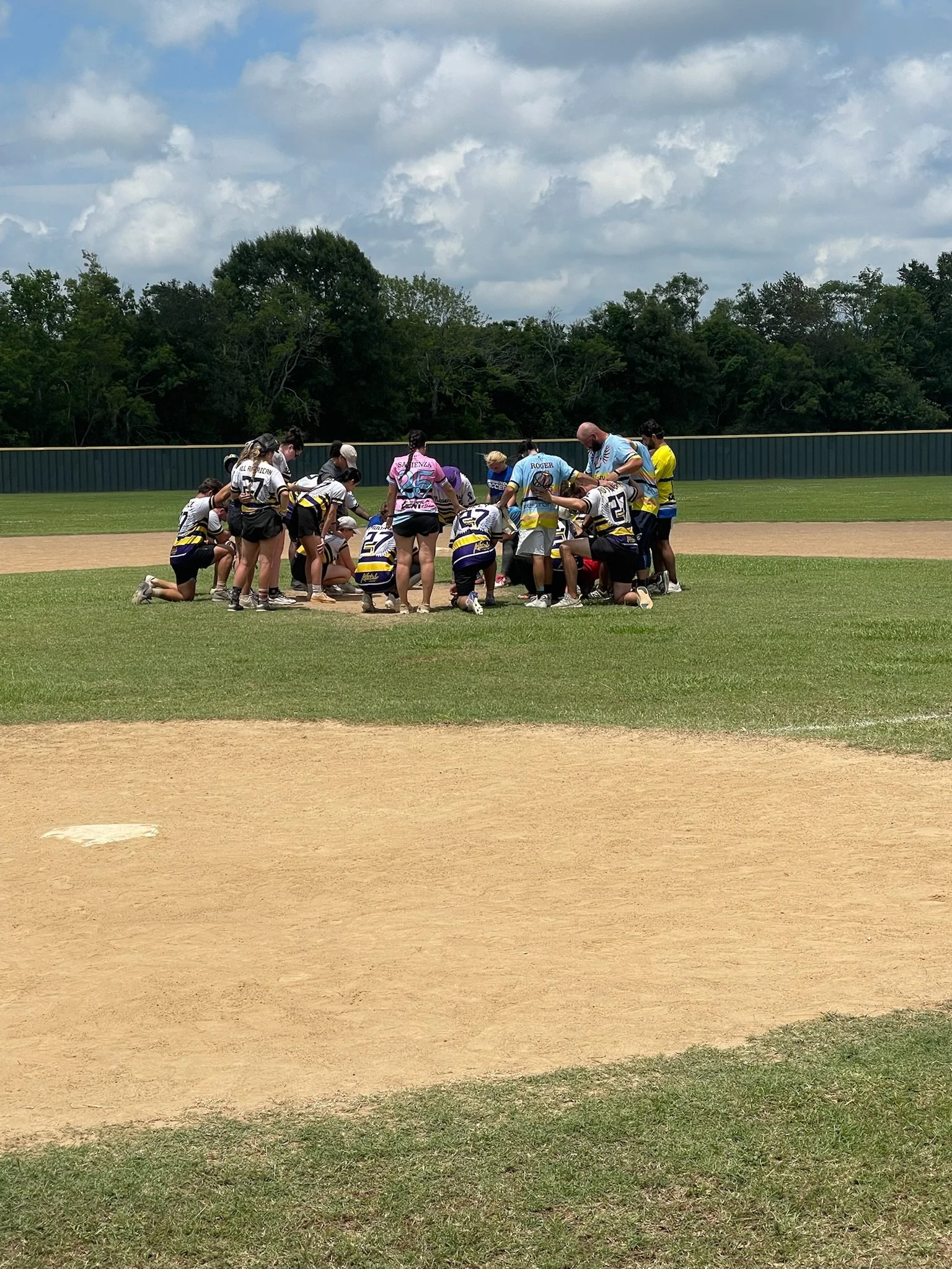A group of people, likely a softball team, gathered on a baseball field practicing or listening to instructions, with lush green trees and a cloudy sky in the background.