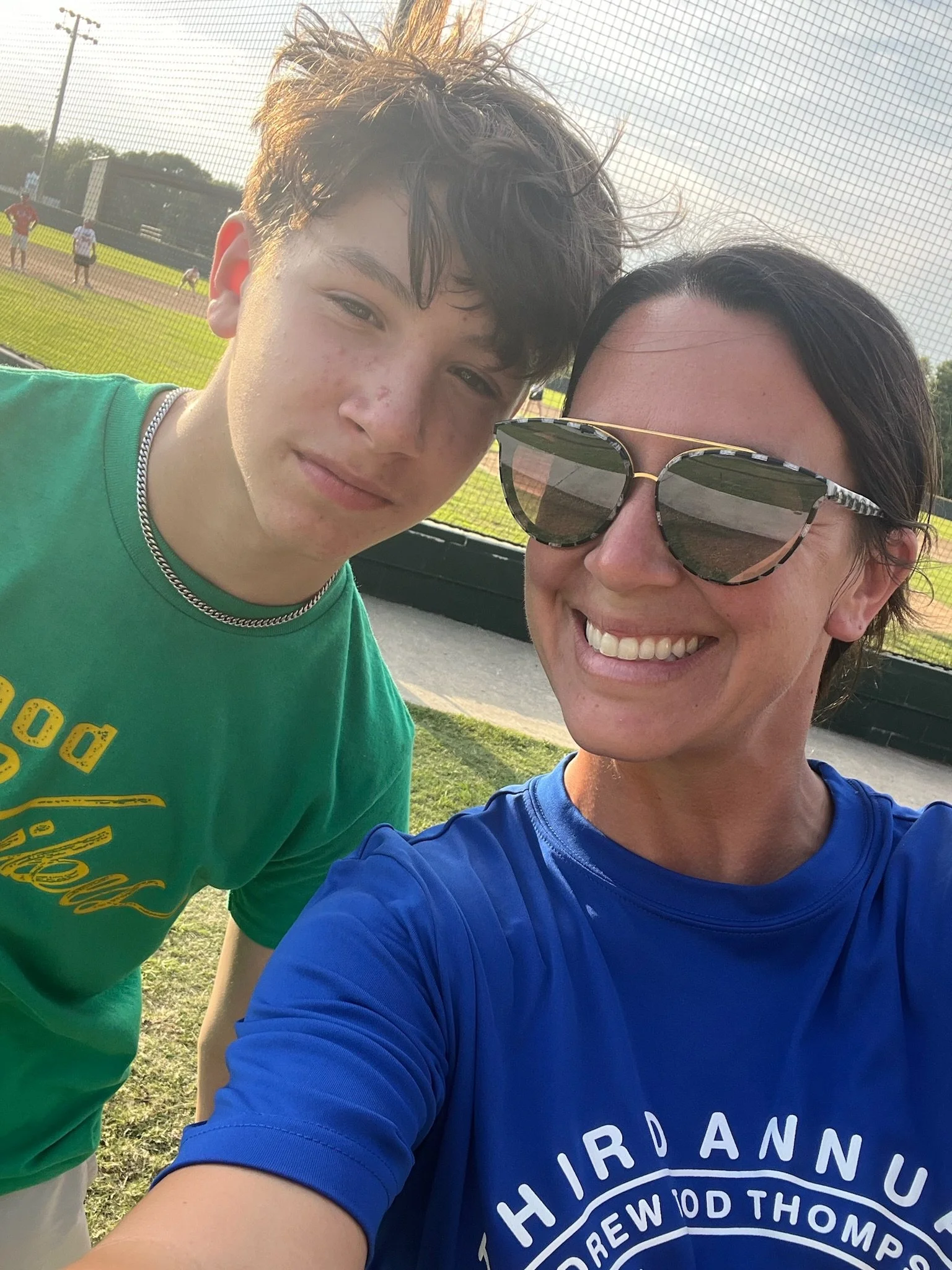 A young man and a woman taking a selfie at a baseball field during a sunny day. The young man has curly brown hair, wearing a green t-shirt and a silver chain necklace. The woman is wearing sunglasses with a patterned frame and a blue shirt.