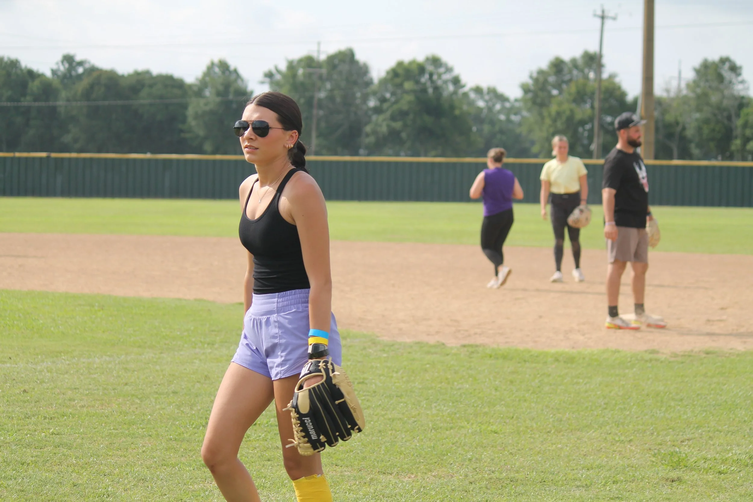 A woman in sunglasses, a black tank top, and purple shorts standing on a grass field holding a baseball glove, with three people in the background on a baseball field.