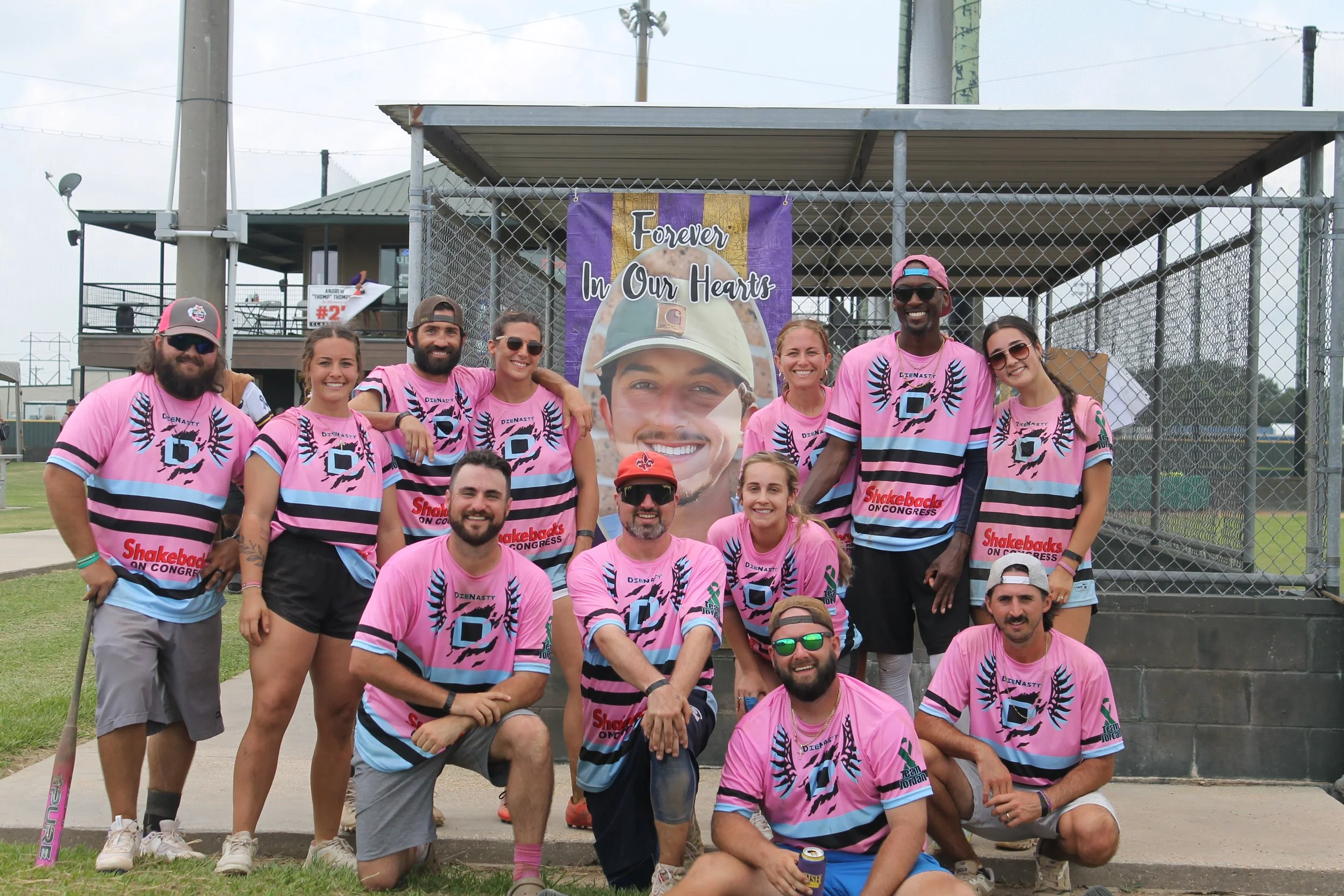 Group of people wearing matching pink and blue sports jerseys posing outdoors in front of a chain-link fence and a banner that reads 'Forever in Our Hearts' with a photo of a young man wearing a baseball cap.