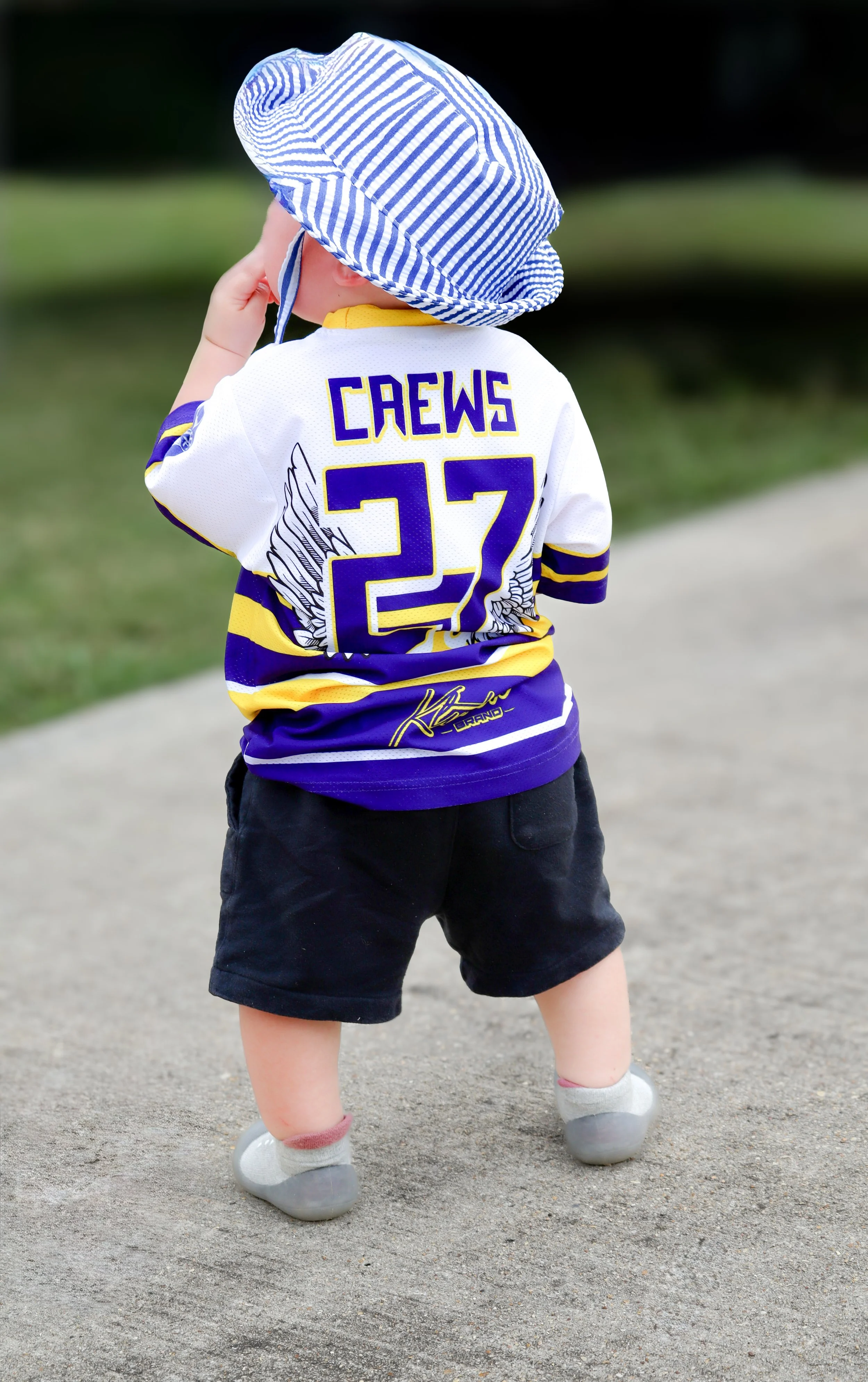 Back view of a young child wearing a striped sun hat, a hockey jersey with the name 'Craws' and number 27, and black shorts, standing on a sidewalk.