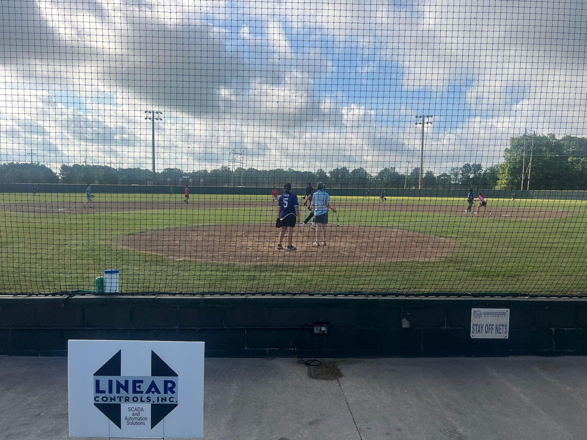 View of a baseball game from behind a safety net, showing players on the field and on the bases, with a partly cloudy sky and power lines in the background.