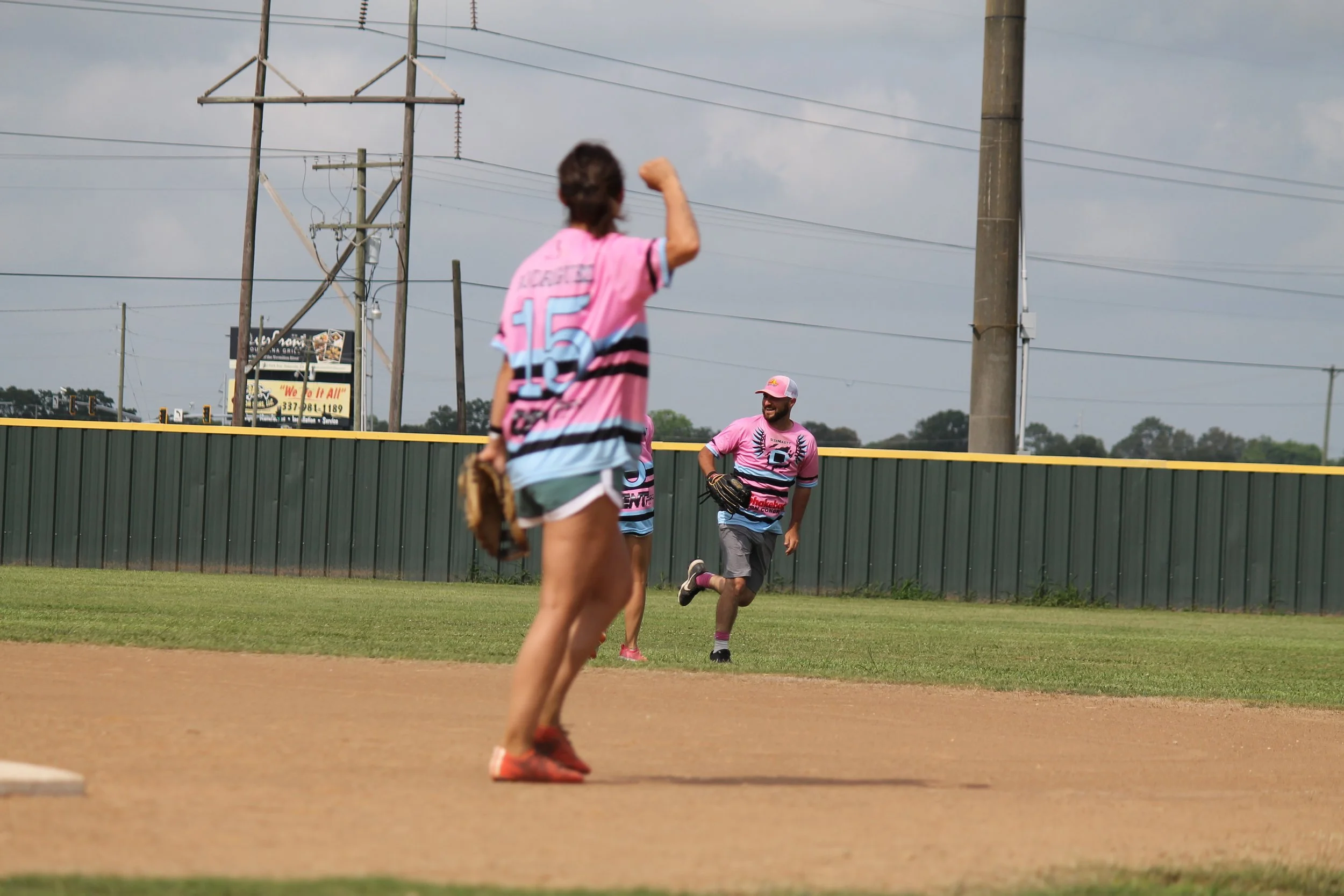 A baseball player ready to catch a ball with two teammates running toward him on the field, all wearing pink and black jerseys, during daytime with cloudy sky.