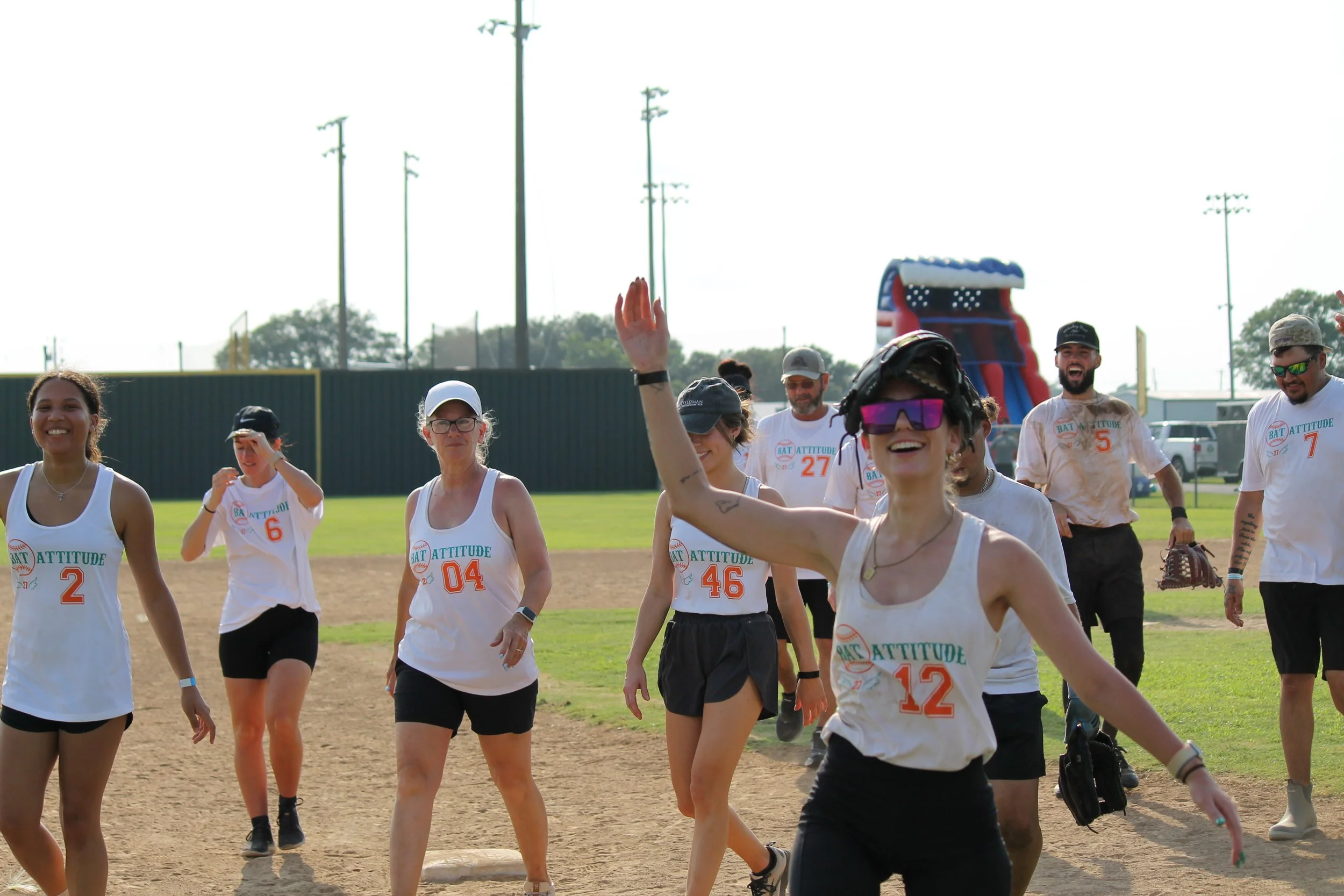 Group of diverse people in athletic clothing on a baseball field, smiling and waving, with some in sunglasses, and a water slide in the background.