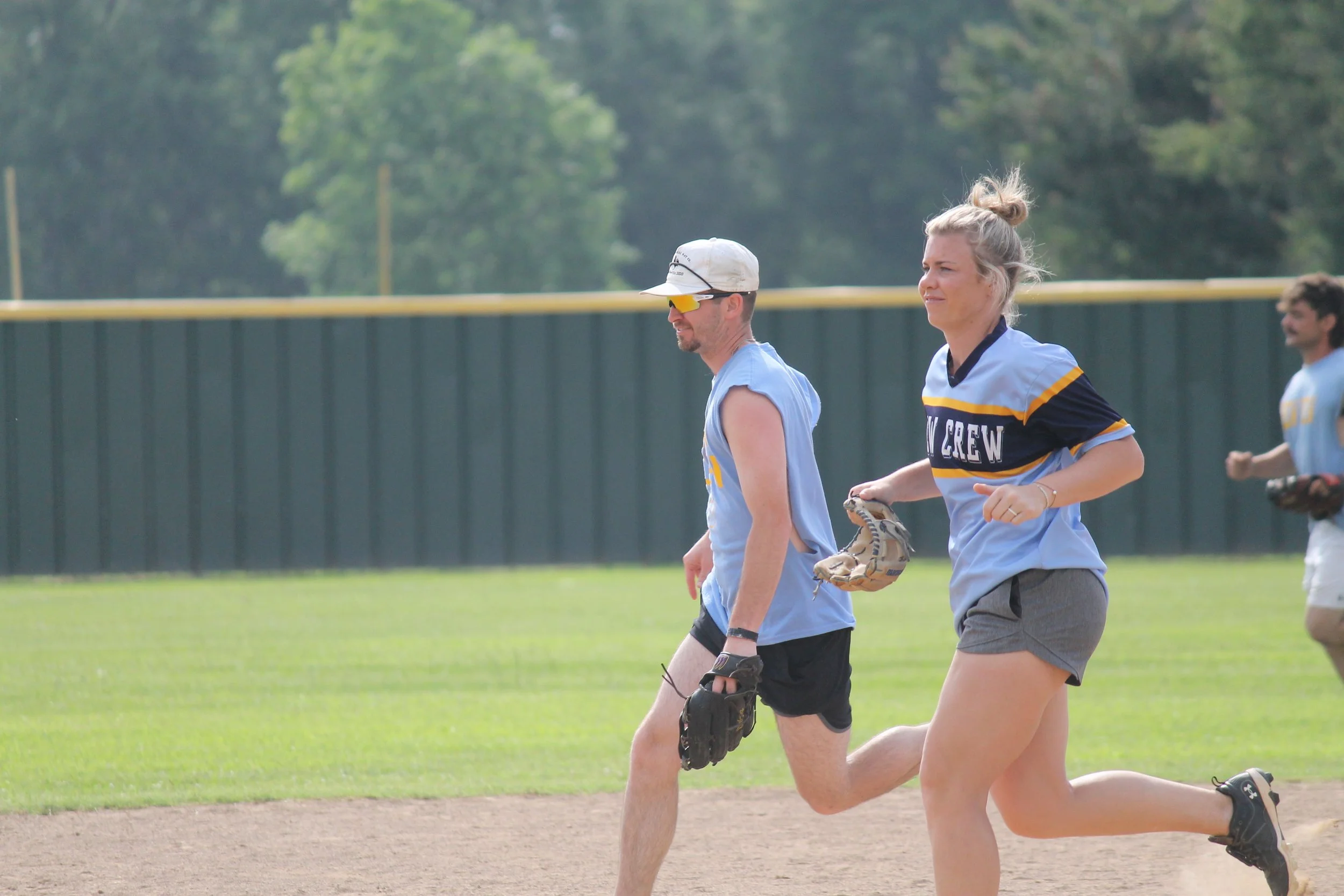 A group of young people playing baseball on a grassy field, with a person holding a baseball glove in the foreground and a green fence and trees in the background.