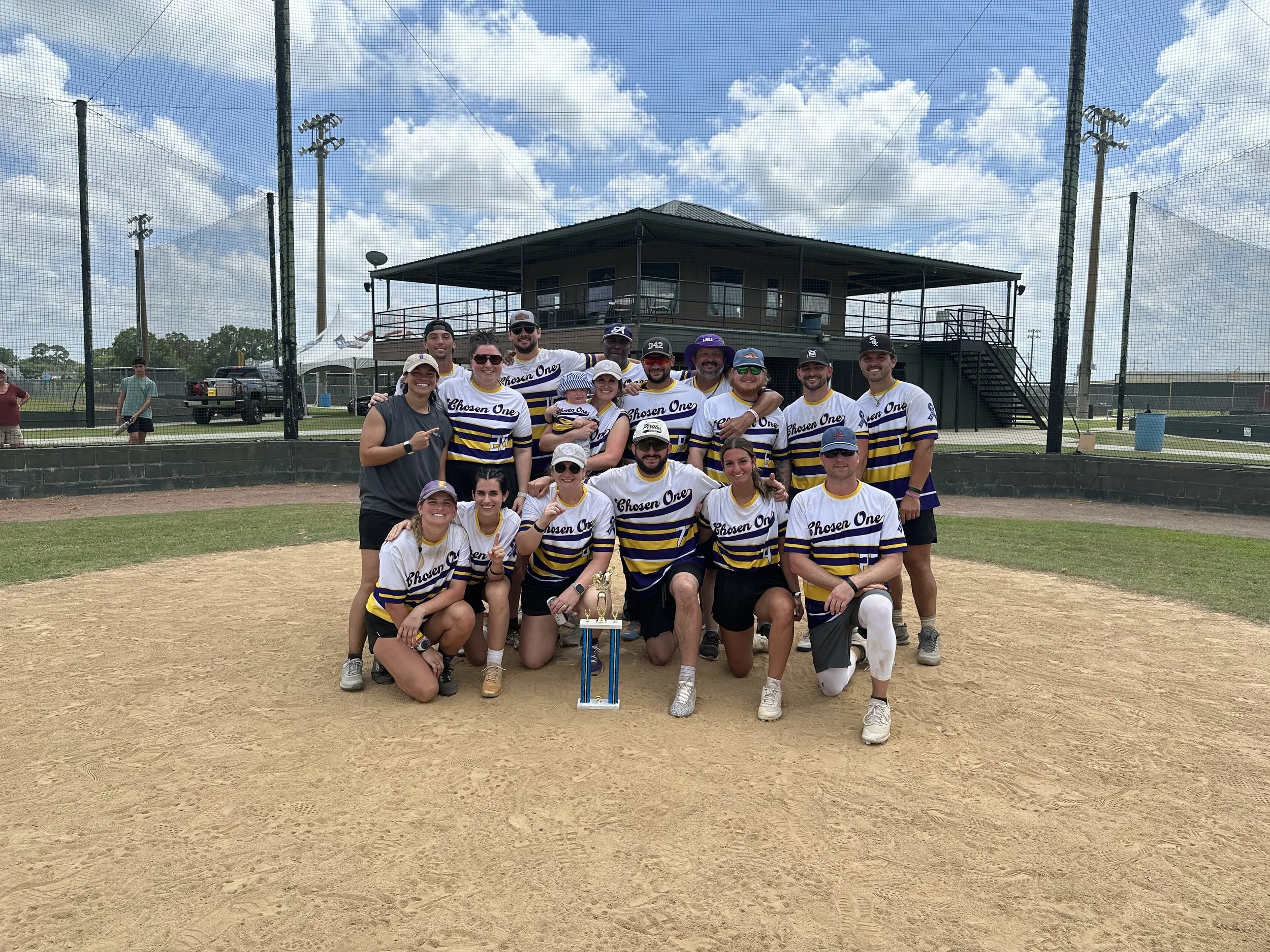 Group of people on a baseball field, celebrating a victory, with a trophy in front. They are wearing matching jerseys that say 'Chosen One' and are smiling at the camera.