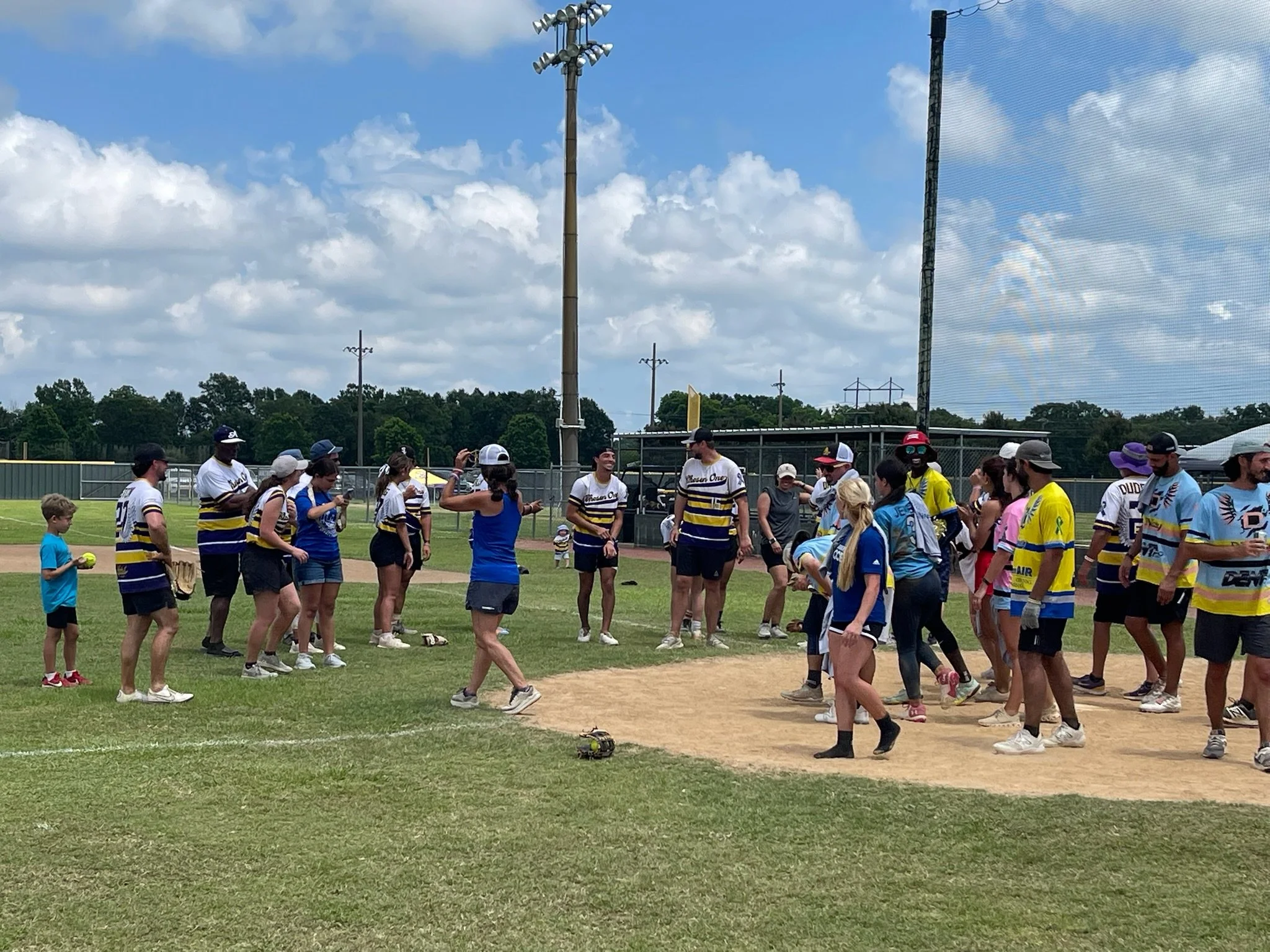 People gathered on a baseball field, some in sports uniforms, with a clear sky and clouds overhead.