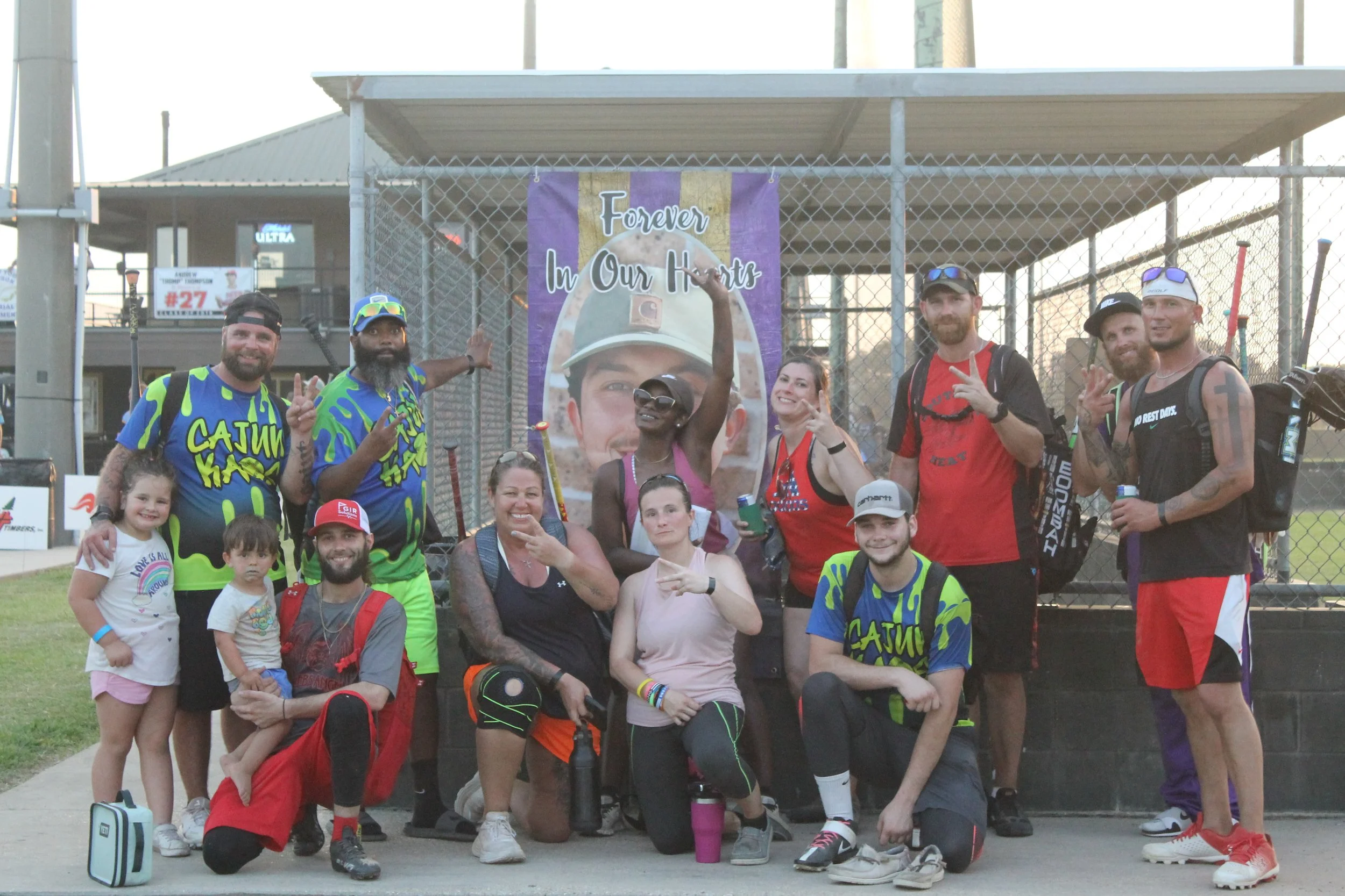 Group of people posing in front of a chain-link fence with a banner that reads "Forever in Our Hearts" and shows a young man in a helmet. The group includes adults, children, and teenagers, some wearing sports jerseys and casual clothing, and they ar