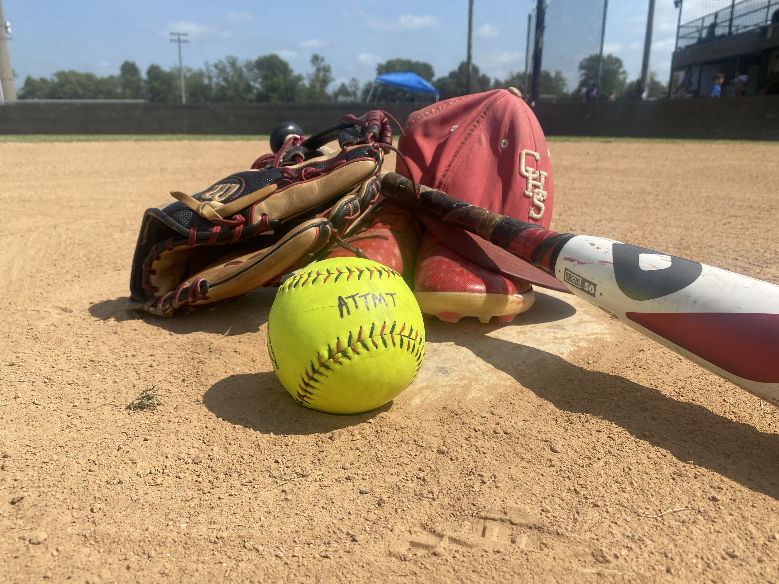 Softball glove, yellow softball, baseball bat, pink baseball bag on a dirt baseball field.