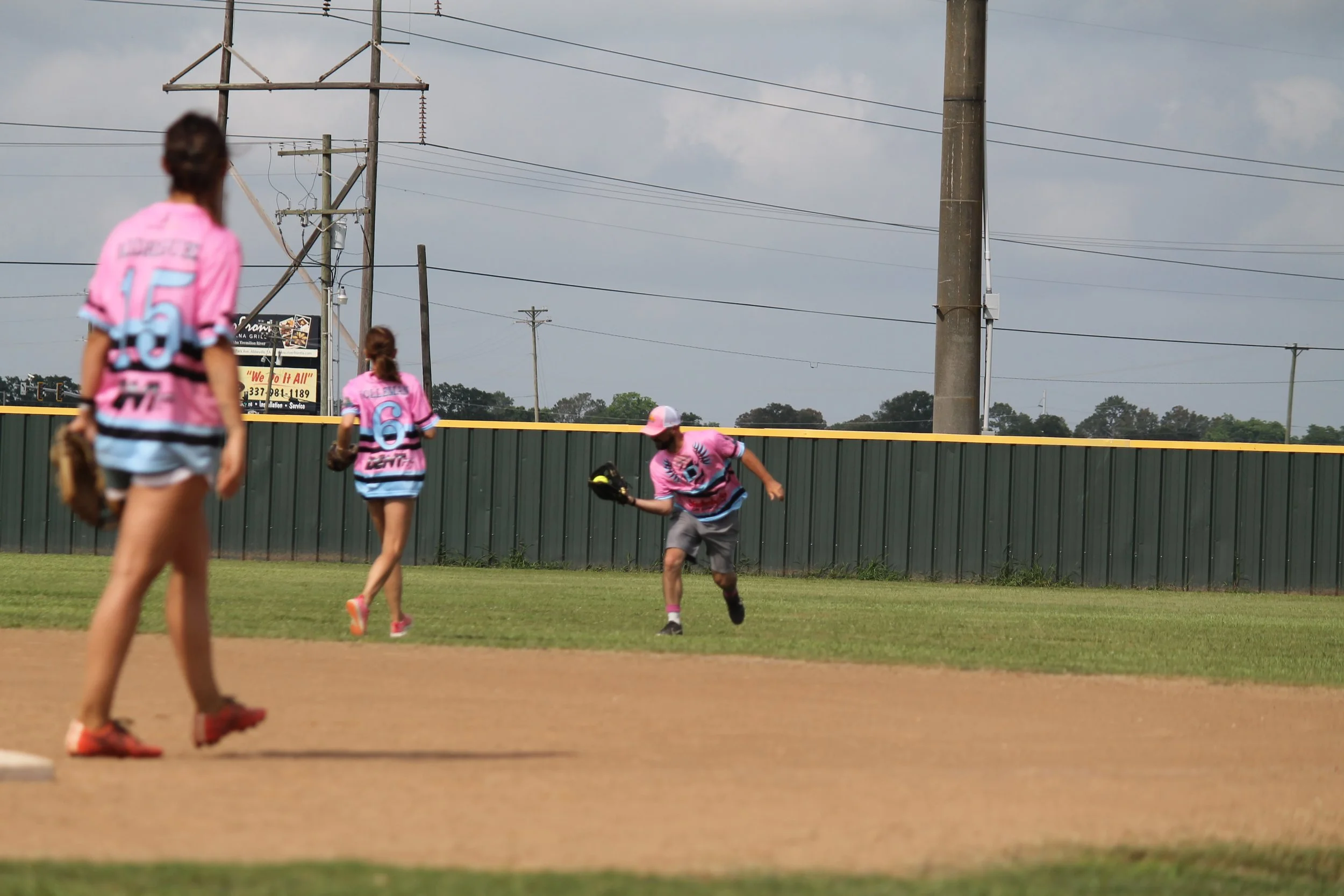 Three young baseball players in pink jerseys on a baseball field, with one player running while holding a glove and cap, and two others standing nearby, during daytime with cloudy sky and power lines in the background.