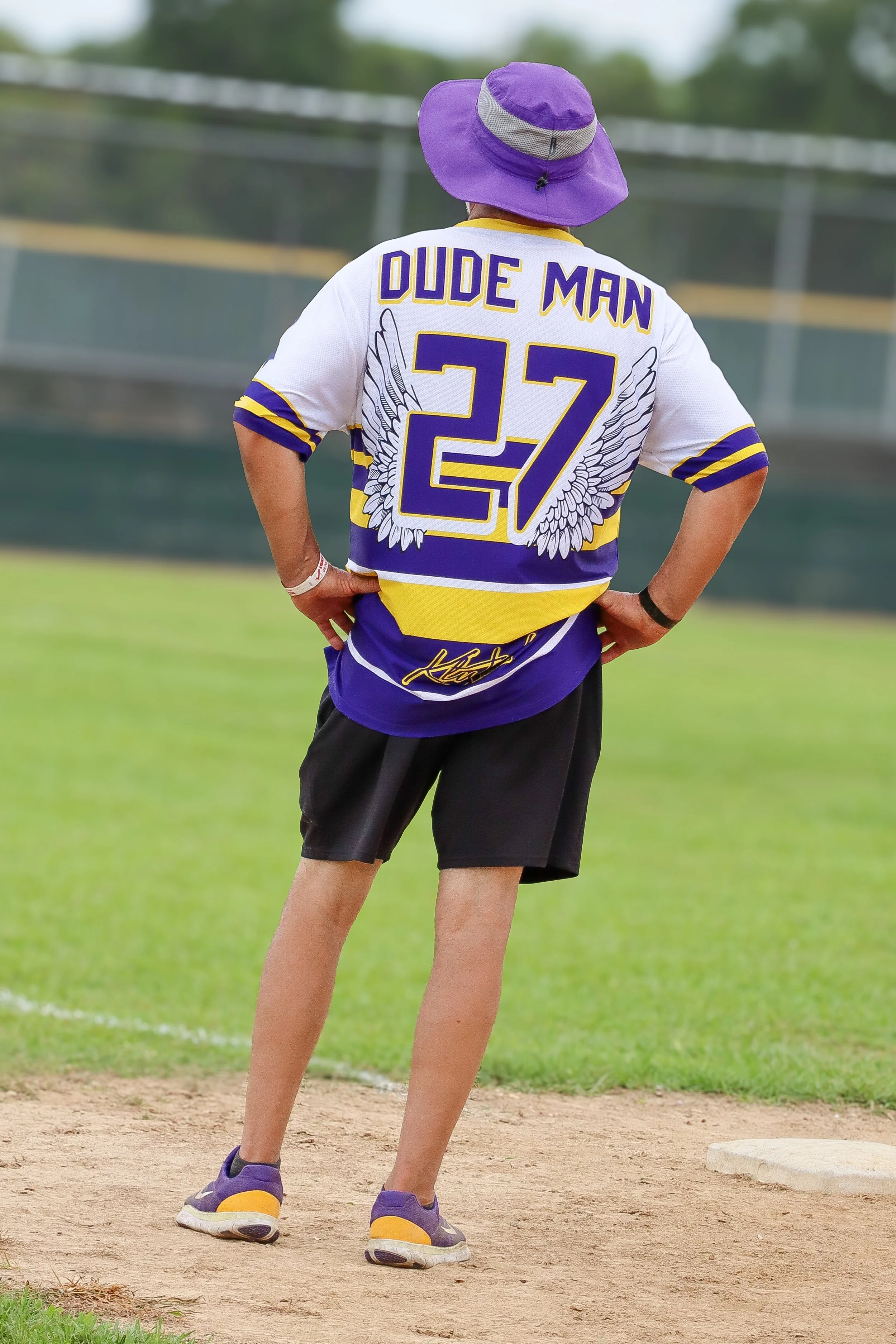 A person wearing a purple and yellow sports jersey with the name 'Dude Man' and the number '27' on the back, standing on a baseball field with hands on hips and a purple hat.