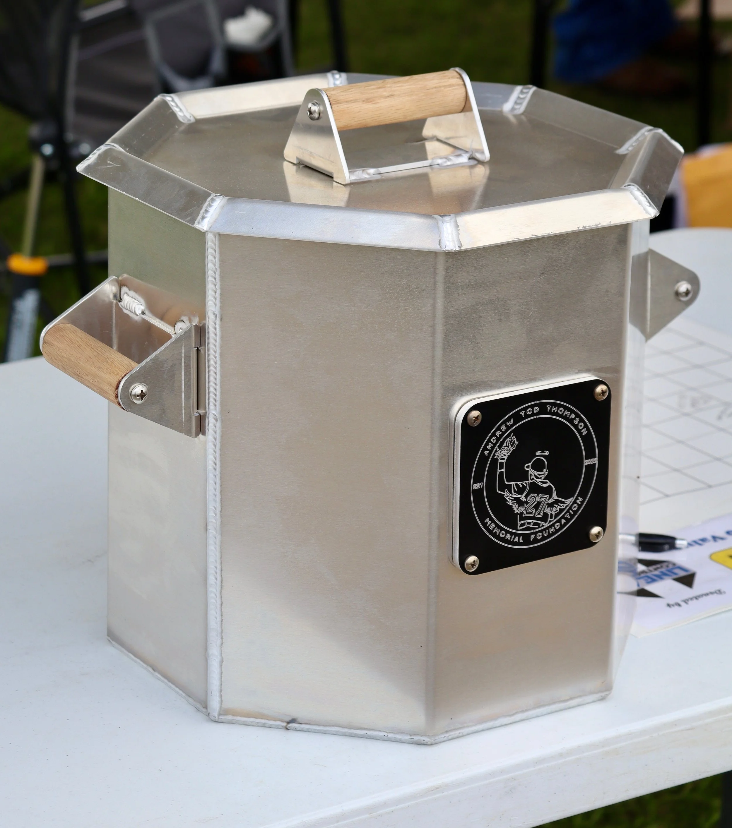 A metal conical-shaped object with wooden handles on each side and on top, resting on a white table outdoors.