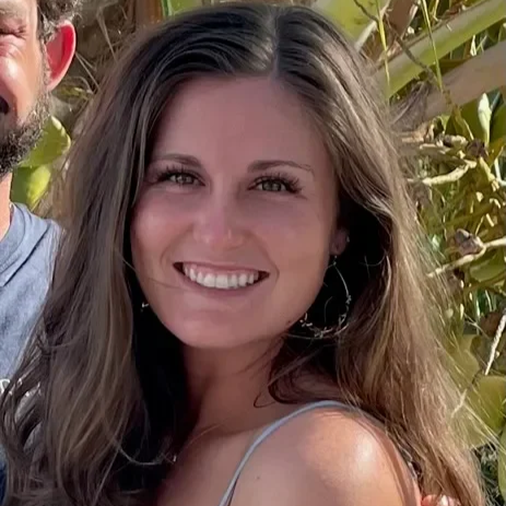 A young woman with long, wavy brown hair smiling outdoors in front of greenery.