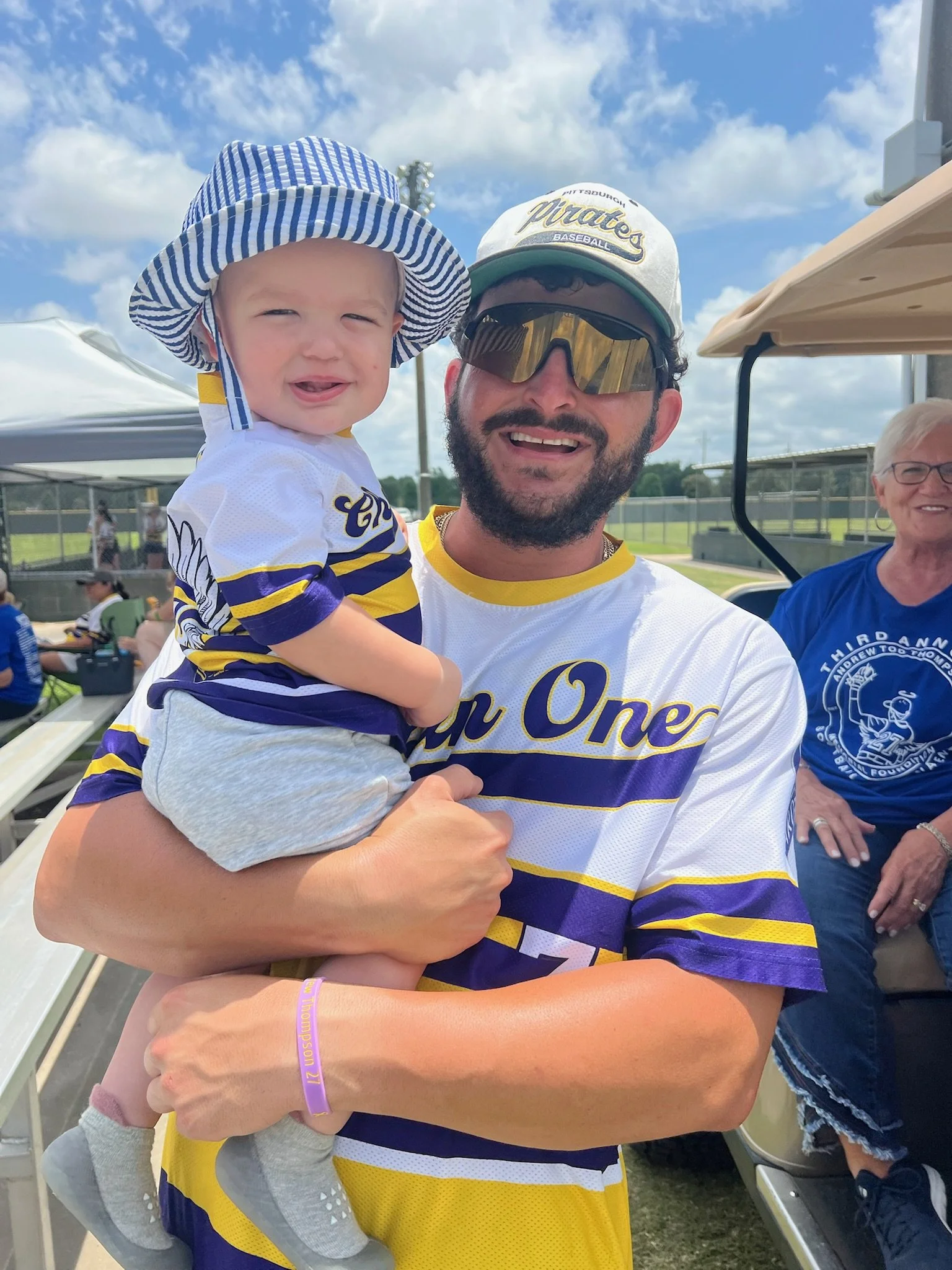 A man holding a young child at a baseball game, both wearing Pittsburgh Pirates jerseys. The man is wearing sunglasses and a baseball cap, and they are smiling under a partly cloudy sky.