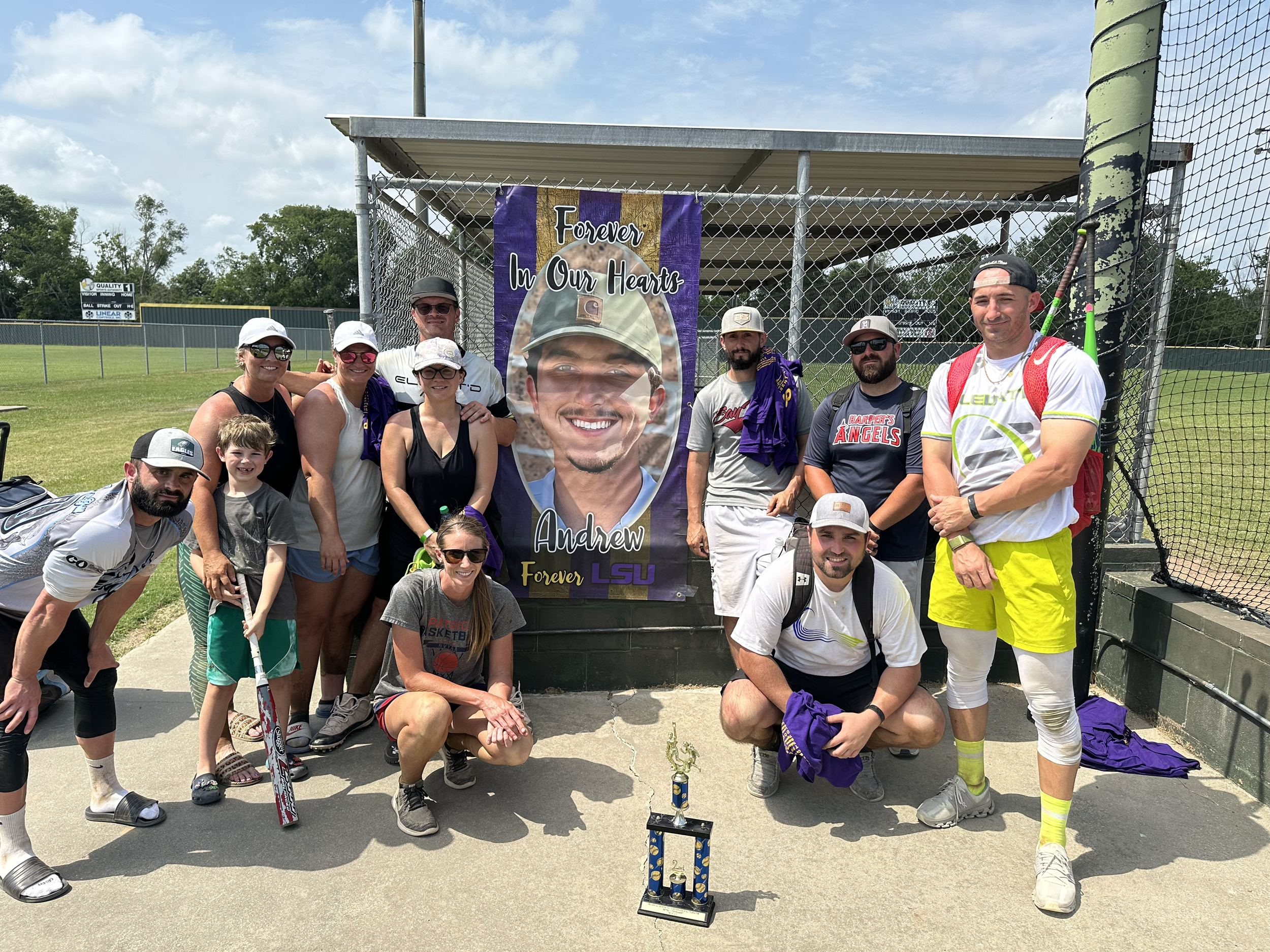A group of people posing in front of a memorial for Andrew, who played baseball at LSU. The group includes adults and children, some holding baseball equipment. A trophy is placed on the ground in front of them. The background features a baseball fie