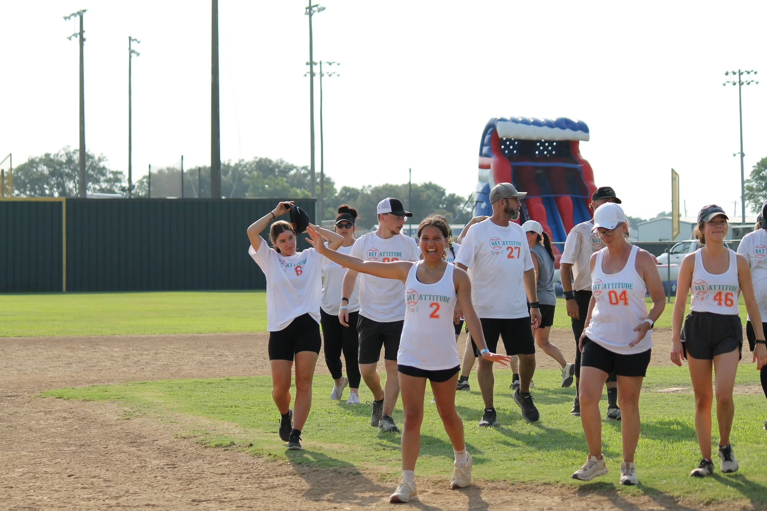 Group of young women and men on a baseball field, wearing athletic clothes with the words 'BAT ATTITUDE' and numbers, walking and smiling, with a large inflatable slide in the background.